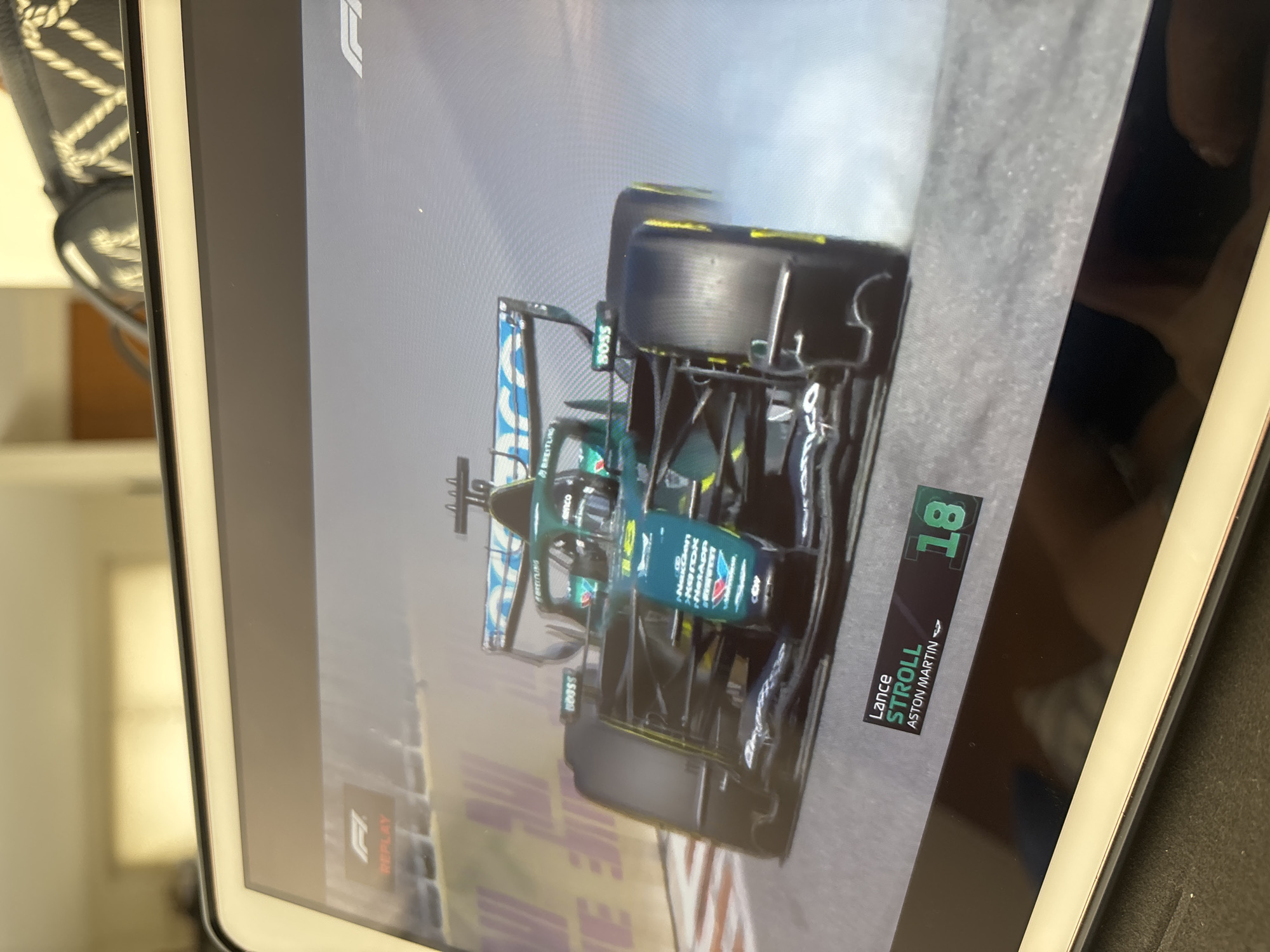 The image shows a close-up view of a screen displaying Formula 1 footage. The focus is on the Aston Martin race car, driven by Lance Stroll (number 18), during a replay. The car is shown amidst smoke or mist. Details visible on the car include sponsor logos such as "BOSS", "Breitling", "NetApp", and "Naken." The footage is displayed on a tablet device, and part of a shoe is visible in the upper left corner of the frame. The setting appears to be indoors, with a blurry background suggesting a room interior. The information provided does not include any location in Puebla, Mexico.
