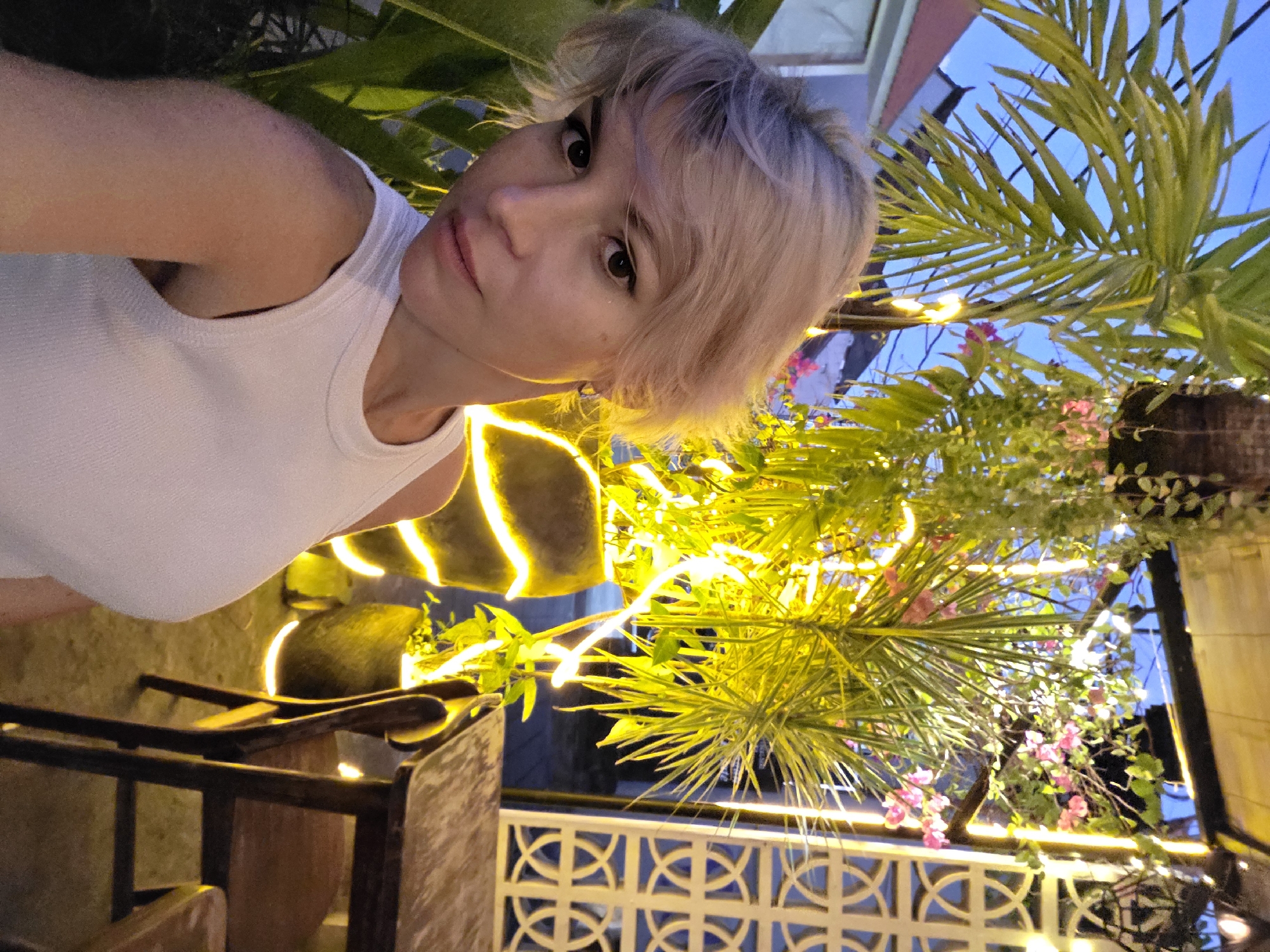 A woman with short, light-colored hair is taking a selfie outdoors in what appears to be a tropical setting at dusk. She is wearing a white tank top and looking directly at the camera with a neutral expression. Her left arm and shoulder are visible, along with part of her chest. The background features lush green palm fronds, some of which are illuminated by bright yellow strip lights. Small pink flowers are interspersed among the foliage, adding pops of color. In the lower portion of the image, there are glimpses of dark wooden furniture and a white decorative railing with a geometric pattern. The sky above is a deep blue, indicative of twilight. The lighting suggests it is evening. The environment seems to be an outdoor dining area or lounge, possibly a restaurant or resort, given the decorative elements and the presence of seating. The overall mood is relaxed and perhaps slightly romantic due to the ambient lighting. No text is visible in the image.