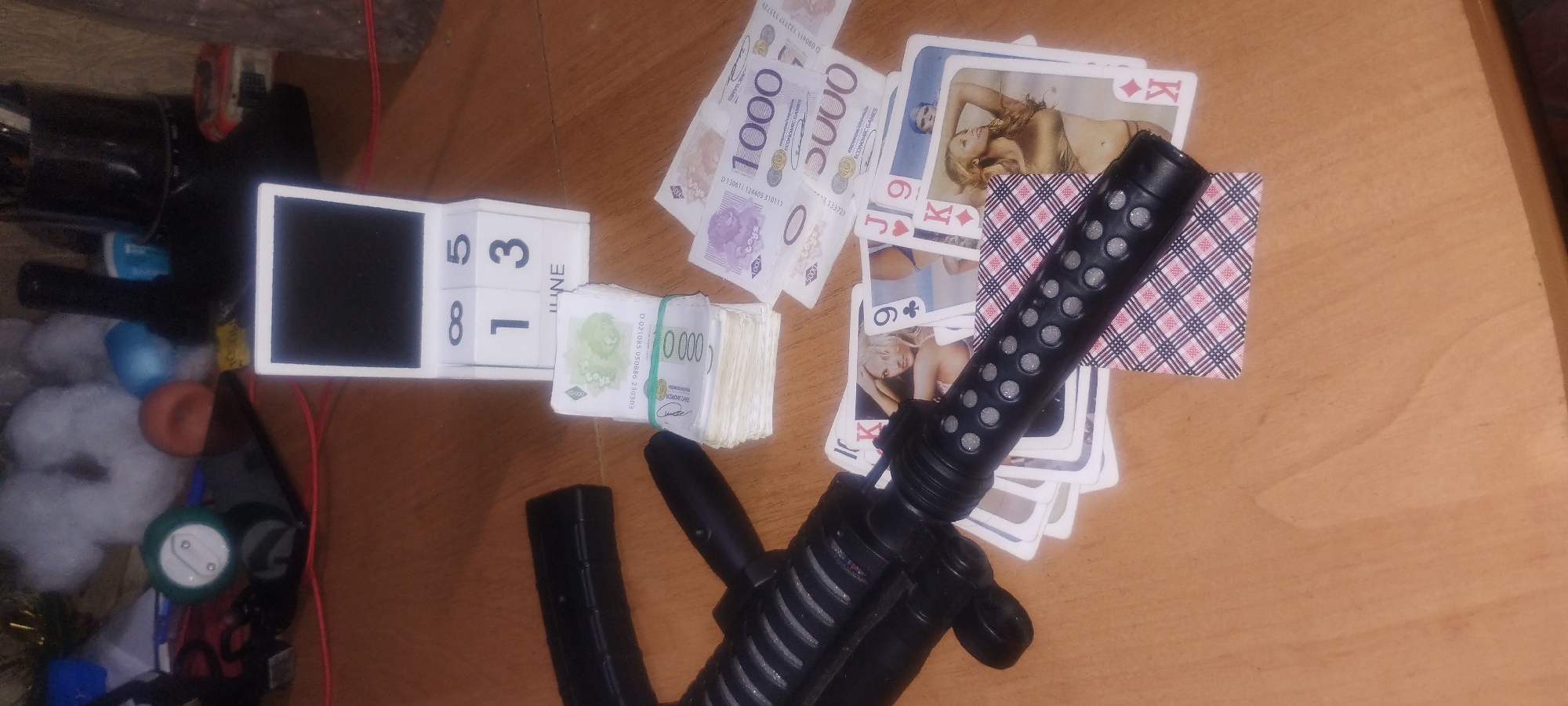 The image shows a collection of items on a wooden surface, possibly a table. The items include a stack of banknotes, several playing cards, a black object resembling a firearm or a part of one, and a white perpetual calendar displaying the date "13 JUNE". The banknotes are banded together and appear to be of various denominations. The playing cards have images of women on them and are fanned out. The black object has a textured grip and appears to be a realistic replica or toy. The perpetual calendar is a cube with numbers and the word "JUNE" printed on its faces. The background is out of focus, but suggests a cluttered environment with various objects and lights. The scene does not provide any information about a specific location in Zaporizhzhia, Ukraine, other than it is indoors on a wooden surface.