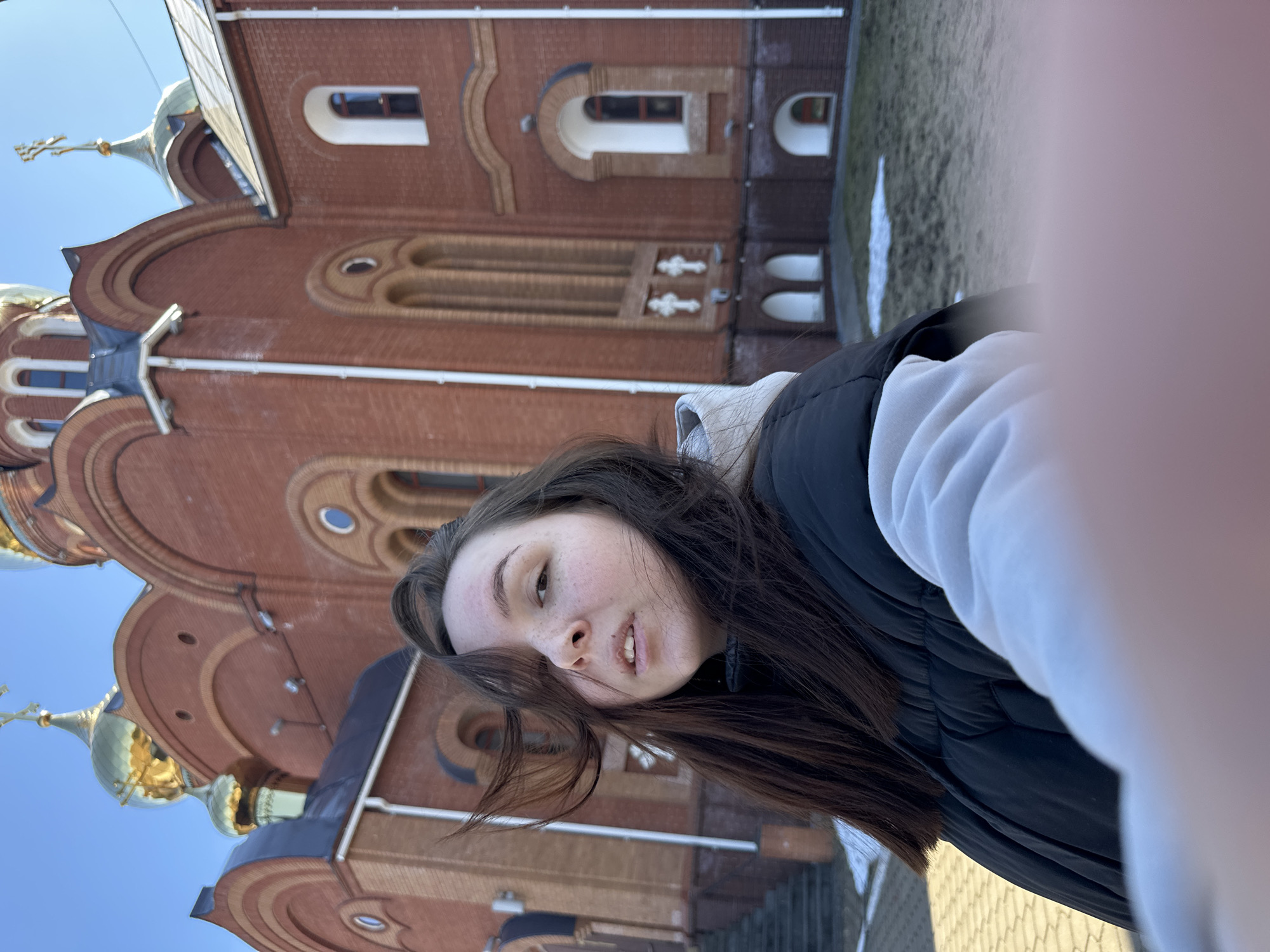 A woman with freckles and dark hair is standing in front of the Church of the Holy Great Martyr George the Victorious in Chaikovsky, Russia. She is wearing a black vest over a white hoodie. The church is a red brick building with golden domes and crosses on top. There is some snow on the ground. The photo is taken at an angle.