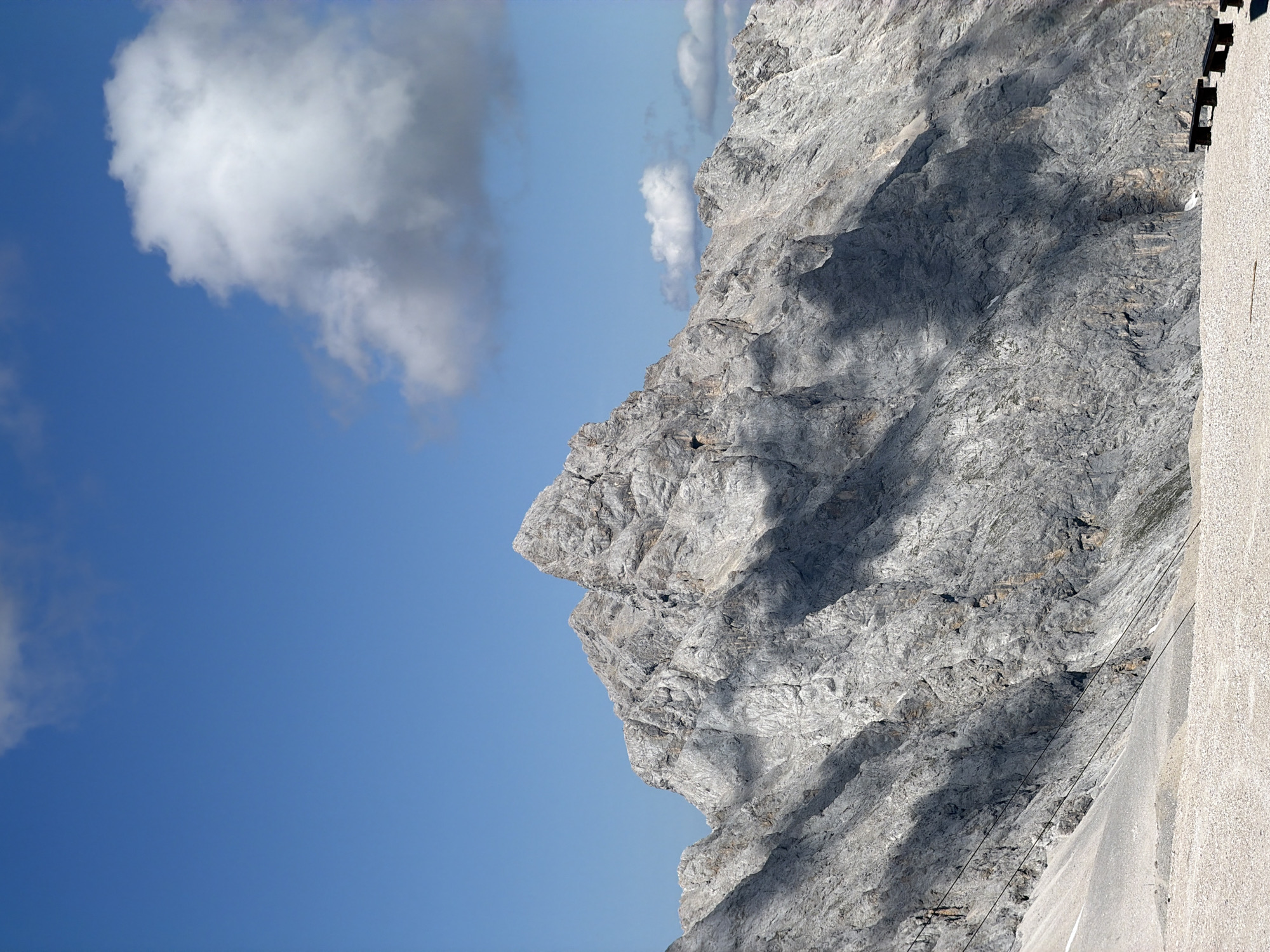 This is a vertical, medium shot of the Zugspitze mountain, the highest peak in Germany, located near Garmisch-Partenkirchen. The mountain is composed of rough, grey rock and is illuminated by sunlight, casting sharp shadows across its slopes. The sky is a vibrant blue, with a few white and grey clouds visible. In the foreground, the ground is covered with light-colored gravel and stones. A dark-colored bench is partially visible on the right side of the frame. There are no people in the image.