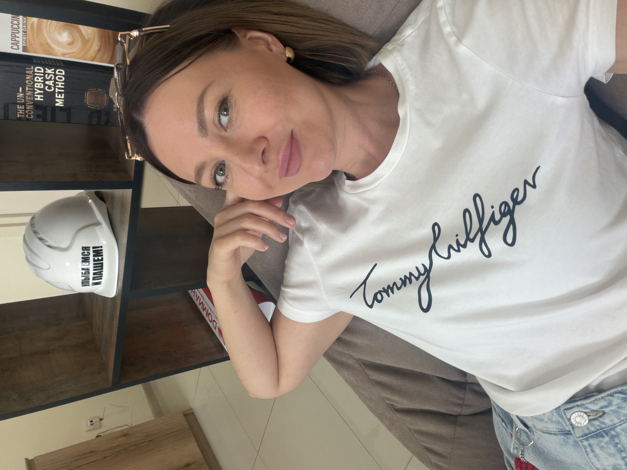 A woman is pictured lying down indoors, looking calmly towards the camera. She has light brown hair, green eyes, and is wearing a white short-sleeved t-shirt with "Tommy Hilfiger" written across the chest in black cursive. A gold chain is visible around her neck, and she has a small gold earring in her right ear. Her left hand is resting near her face. She appears to be on a sofa or cushioned surface.
Behind her is a dark wooden bookshelf containing several items. Visible book titles include "CAPPUCCIN SPIRIT DRINK" and "THE UN-CONVENTIONAL HYBRID CASK METHOD". On a lower shelf, a white object, resembling a hard hat or helmet, bears the Russian phrase "УЛЫБАЕМСЯ И ПАШЕМ!" (Ulybayemsya i Pashem!), which translates to "SMILE AND WORK!". Another partially visible item on the shelf shows "DOMMAH" and "СЕТЬ АГЕНТСТВ НЕ..." (Set' Agentstv Ne...), meaning "NETWORK OF AGENCIES". The room has light-colored walls and a power outlet is visible below the shelf. The setting suggests a casual indoor environment, likely a home in Rostov-on-Don, Russia. The time of day appears to be daytime, with soft indoor lighting.