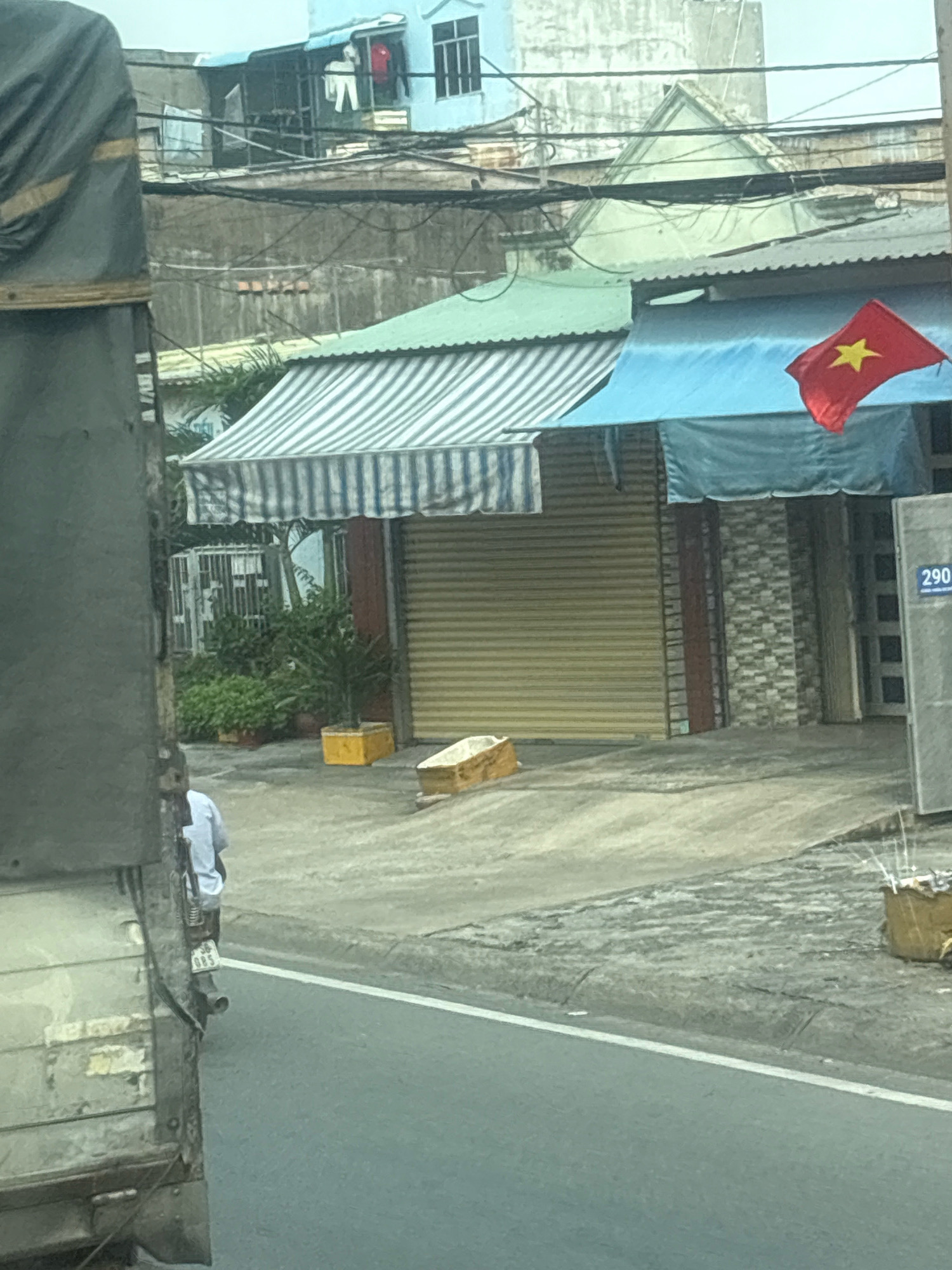 A medium shot captures a street scene in Ho Chi Minh City, Vietnam, with a focus on commercial storefronts and an adjacent street. In the foreground, a large truck with a weathered, grey wooden exterior occupies the left side of the frame, partially obscuring the view. A person wearing a light-colored shirt and dark pants is visible on a motorcycle, facing away from the camera, towards the right side of the image. The motorcycle's license plate, obscured by blur, appears to have the characters "083" visible.
Beyond the truck and the person on the motorcycle, a row of buildings lines the street. The buildings feature awnings, with one prominently displaying blue and white stripes and another a solid blue tarp. A Vietnamese flag, red with a yellow star, is attached to the blue tarp awning, waving in the breeze. The storefronts themselves have metallic roll-up doors, one of which is a light tan color, and another is a darker shade. The building facades vary, with one section displaying a grey and white textured stone pattern. A sign with the numbers "290" is mounted on a grey structure to the right of the buildings.
The ground in front of the buildings is a paved surface, showing some wear and tear. A small, light brown, rectangular object, possibly a box or a small coffin, lies on the ground in front of the tan roll-up door. Greenery, including potted plants and bushes, is visible between some of the storefronts and behind a barred window. Electrical wires are strung overhead, crisscrossing the scene. The sky appears overcast. The overall impression is of a typical urban street with a mix of residential and commercial activity, common in Ho Chi Minh City.