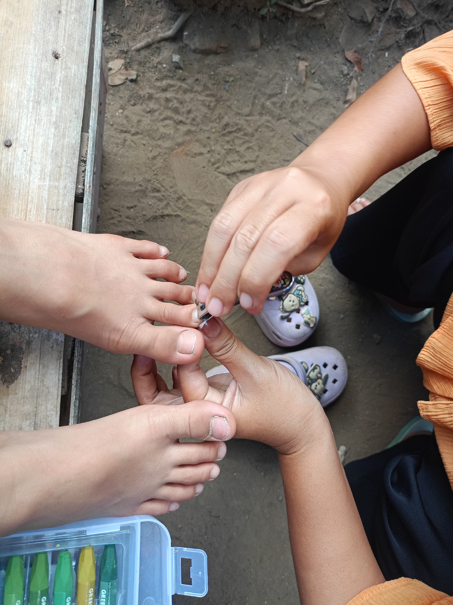 In Jelbuk, Indonesia, a close-up shot captures a person's feet and hands. One person's feet are positioned on a wooden surface, with a small, open container of green and yellow crayons nearby. Another person's hands are actively trimming the toenails of the individual with the exposed feet, using nail clippers. The surrounding ground appears to be dirt and gravel, with some dry leaves scattered about. In the background, light purple clogs adorned with cartoon characters are visible. The overall scene is an outdoor setting, likely a casual, home environment.