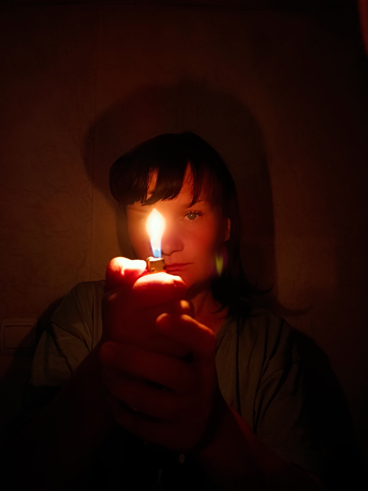 A woman is holding a lit lighter in front of her face. The scene is dark, and the flame of the lighter illuminates her face and casts a shadow on the wall behind her. The location is not discernible in the image.