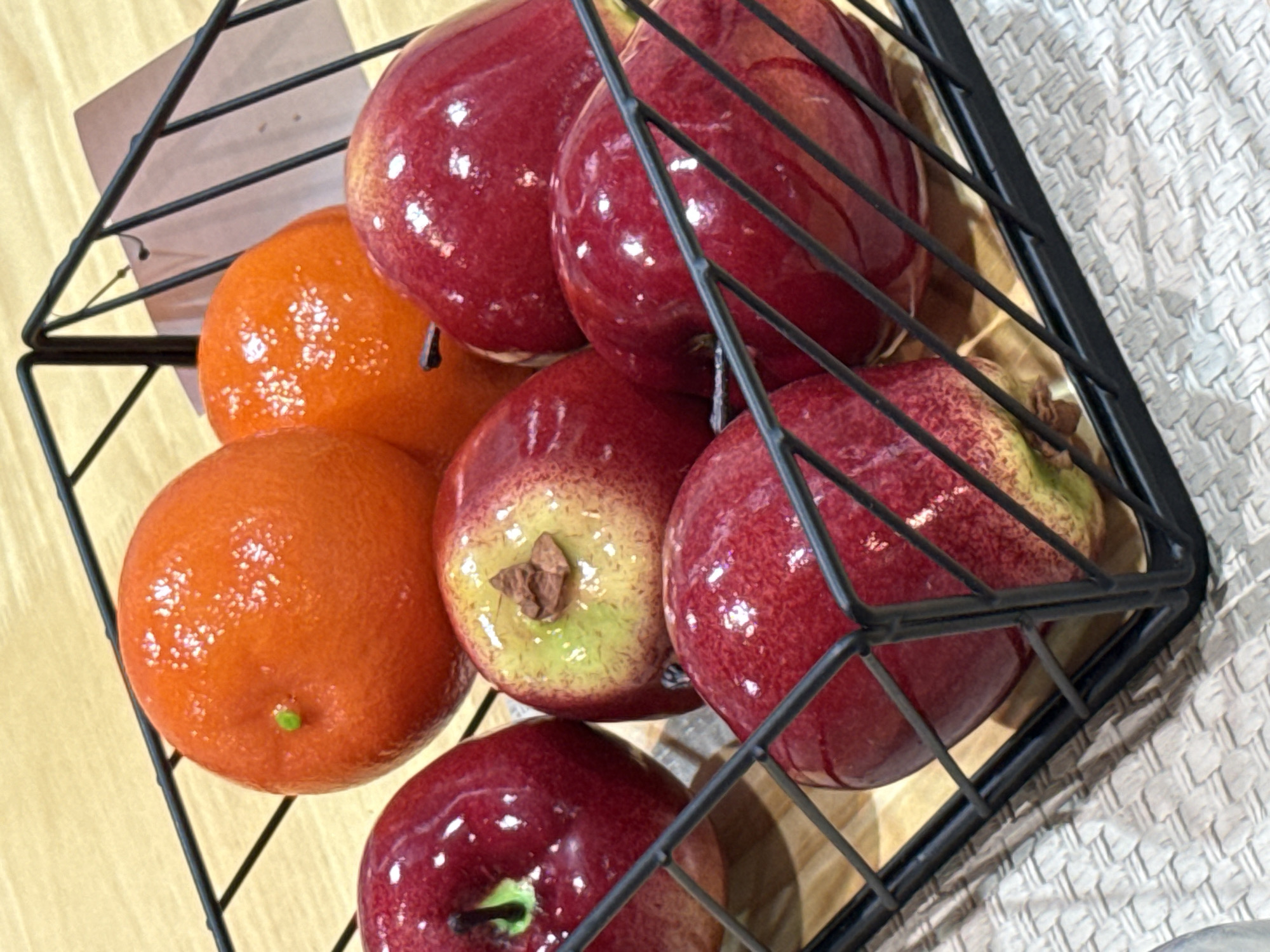 A black wire basket filled with artificial fruits sits on a light-colored woven placemat. The basket contains at least two bright orange, round fruits, possibly tangerines, and at least five shiny, deep red apples. The apples have green and yellow undertones near their stems. The background shows a light-colored wooden surface. A white tag is attached to the inside of the basket. The scene appears to be an indoor still life. There is no indication of location in Baan Chaweng Noi, Thailand.