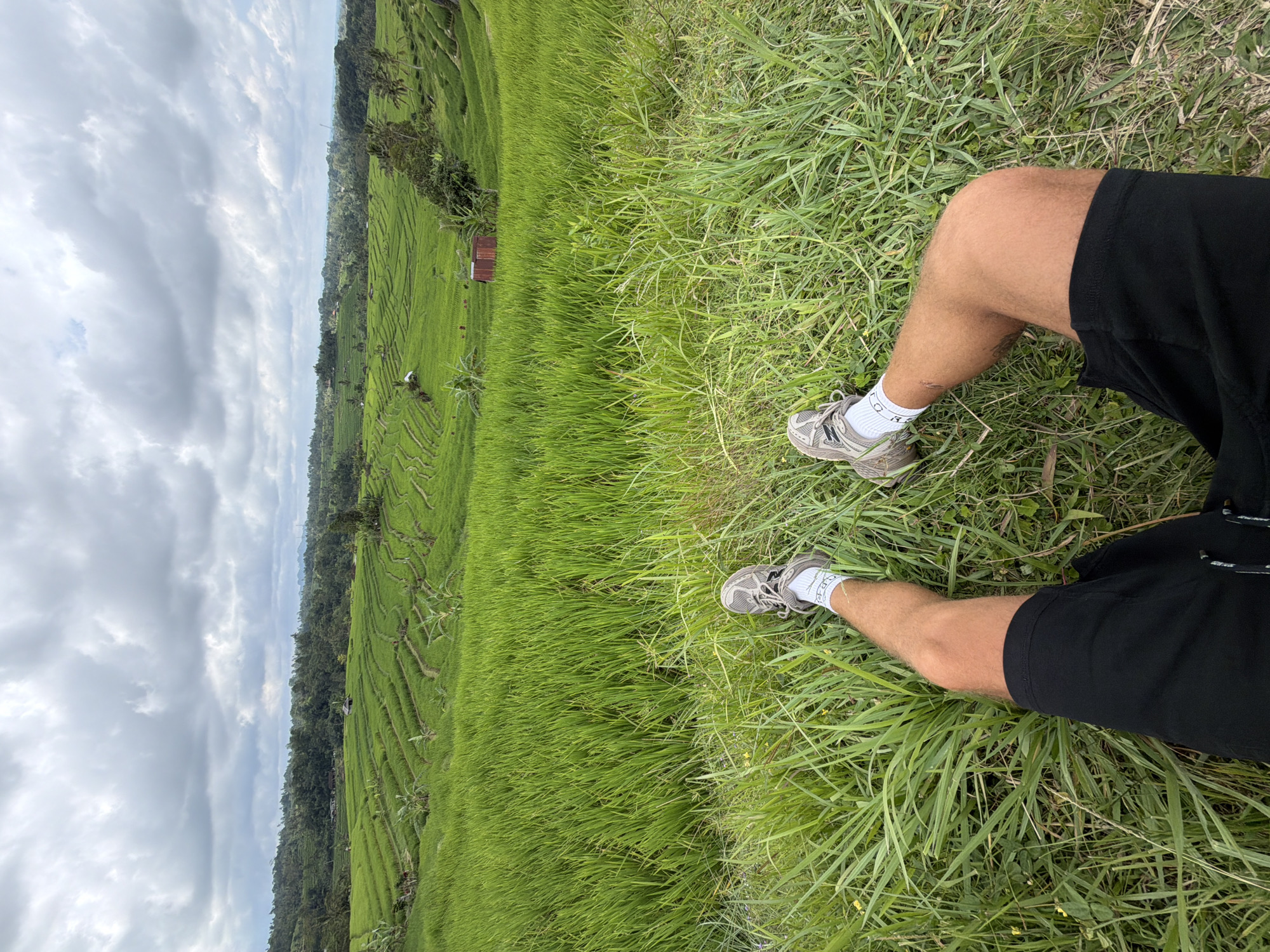 A first-person perspective photograph shows a pair of human legs in black shorts and gray sneakers resting in tall green grass. The background reveals a vast landscape of green rice terraces and lush trees, with a cloudy sky above. A small, reddish-brown building is visible among the terraces. The image suggests a serene and rural setting, with the rice terraces indicating the location to be in Indonesia, likely in a region known for its agricultural landscapes.