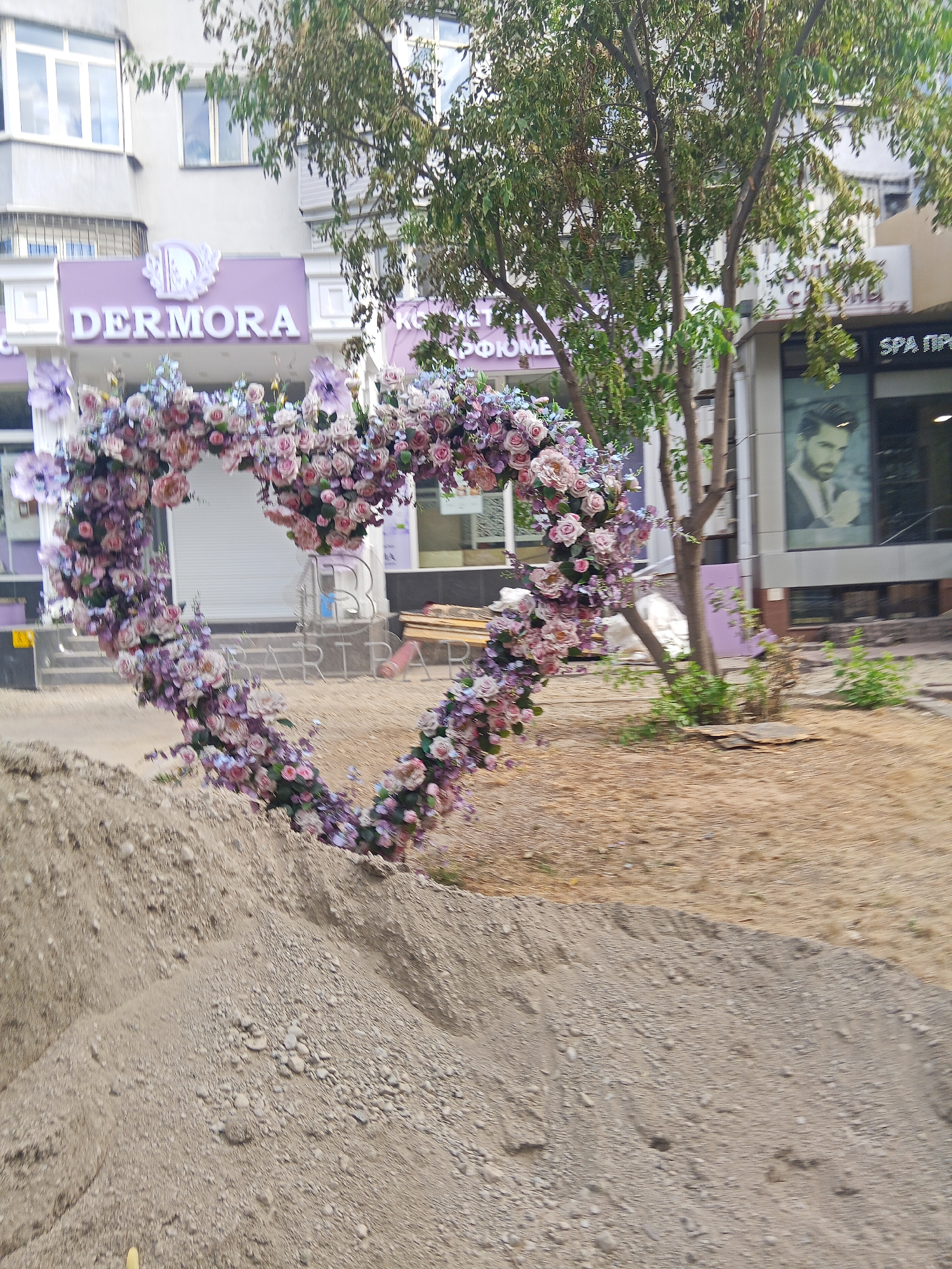 The image depicts a heart-shaped floral archway placed in front of a building with the sign "DERMORA" in purple lettering. The arch is adorned with numerous pink and purple flowers. The building appears to be a commercial establishment, possibly a salon or spa, as suggested by the "SPA" sign visible to the right. The foreground is dominated by piles of sand and gravel, indicating construction or landscaping work. In the background, trees with green foliage are present, and the sky is visible. The overall scene suggests an outdoor setting in a city, likely Almaty, Kazakhstan, given the building's signage and architectural style. No people are clearly visible in the image.