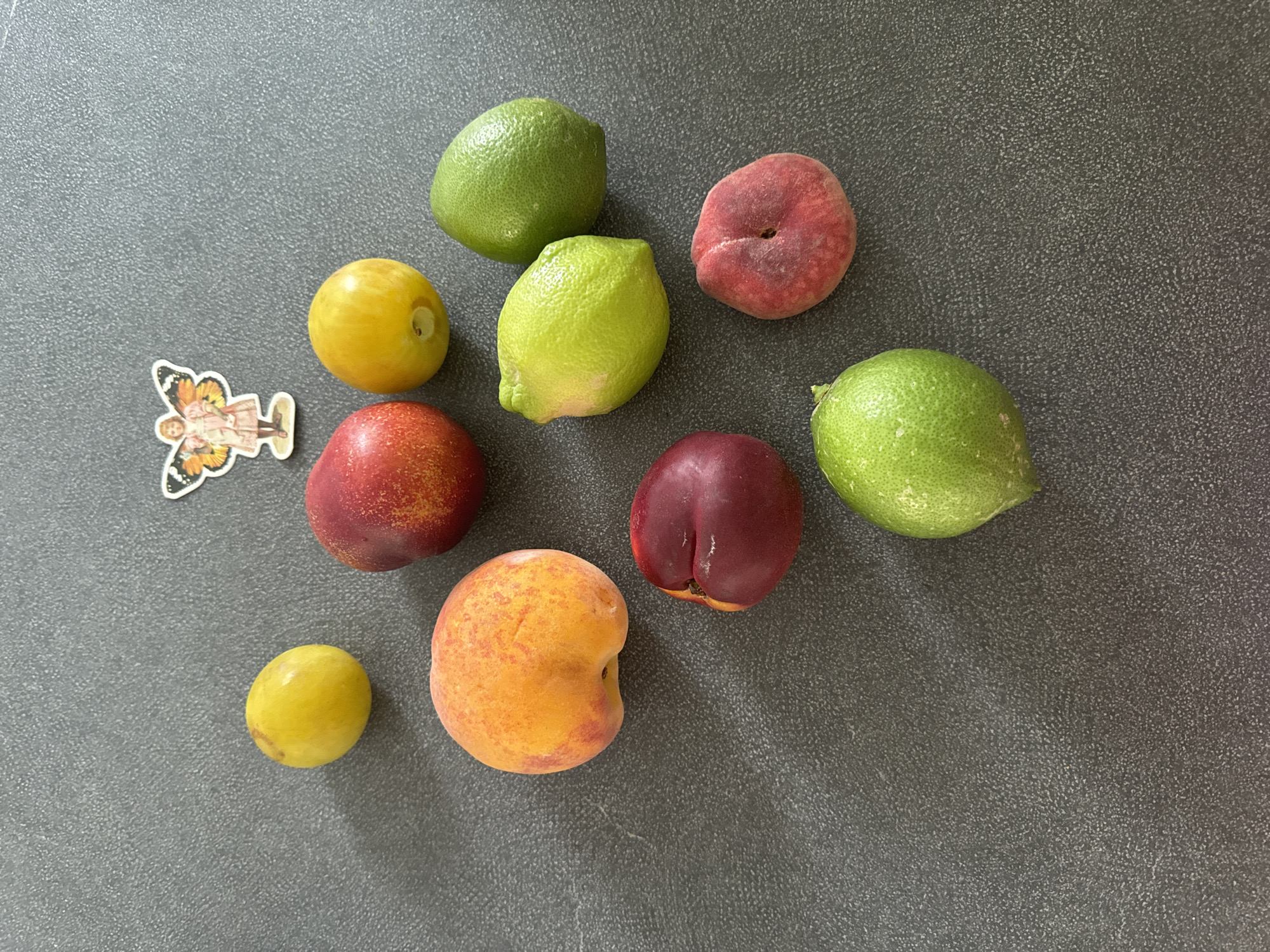 An overhead, close-up shot shows a collection of fruits and a sticker on a dark gray, textured surface. The fruits include two yellow plums or apricots, two red nectarines, one orange and red peach, one flat peach or Saturn peach, and three green limes. The sticker, located near the top center, is of a small figure with butterfly wings. The surface appears to be a countertop or table. No other objects or people are visible, and there are no elements that would indicate a specific location within the city of Murcia, Spain.