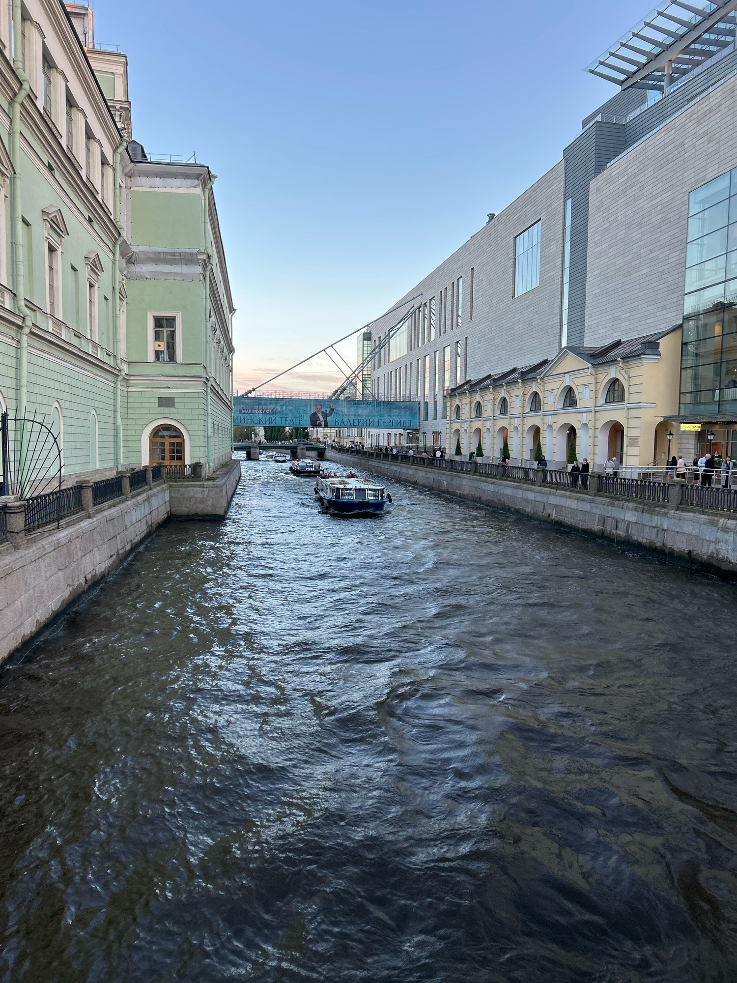 This is a high-angle, eye-level, medium shot of a canal with tour boats. The canal is lined by buildings on both sides. On the left, a pale green building with classical architectural details and arched windows dominates the foreground. On the right, a modern, grey building with a glass facade and a row of yellow, arched storefronts sits along the canal. Several tour boats are visible on the dark, rippling water. A pedestrian bridge spans the canal in the distance, with a banner displaying Russian text. The sky is clear and blue, suggesting it is daytime, likely late afternoon or early evening due to the soft lighting. The overall scene depicts a scenic urban waterway, possibly a tourist area.