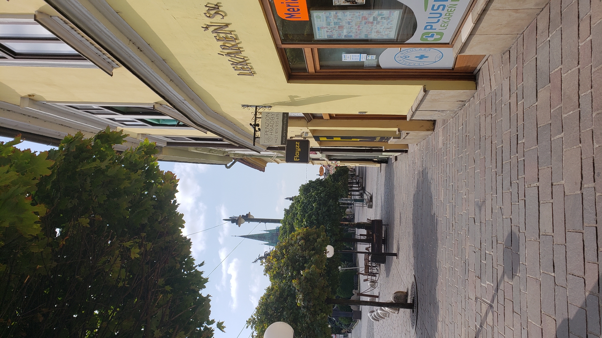 This is a street-level, eye-level, vertical shot of a pedestrian walkway lined with trees and buildings. The walkway is paved with reddish-brown cobblestones and is in the foreground and midground. On the right side of the walkway, a yellow building with large windows and signs is prominently featured. One sign reads "LEKÁREŇ SV. MIKULÁŠA," indicating a pharmacy. Another sign reads "PLUS LEKÁREŇ." On the left side of the walkway, there is a row of evenly spaced trees with rounded canopies, casting shadows on the cobblestones. Beyond the trees, outdoor seating areas with tables and chairs are visible. In the background, a tall church steeple with a green roof pierces the sky. The sky is partly cloudy and bright, suggesting daytime. The overall scene depicts a calm, possibly historic, European town square or street. This is likely a view of the main pedestrian street (Hlavná ulica) in Prešov, Slovakia, given the architectural style and the presence of a pharmacy and a church spire commonly seen in European city centers.