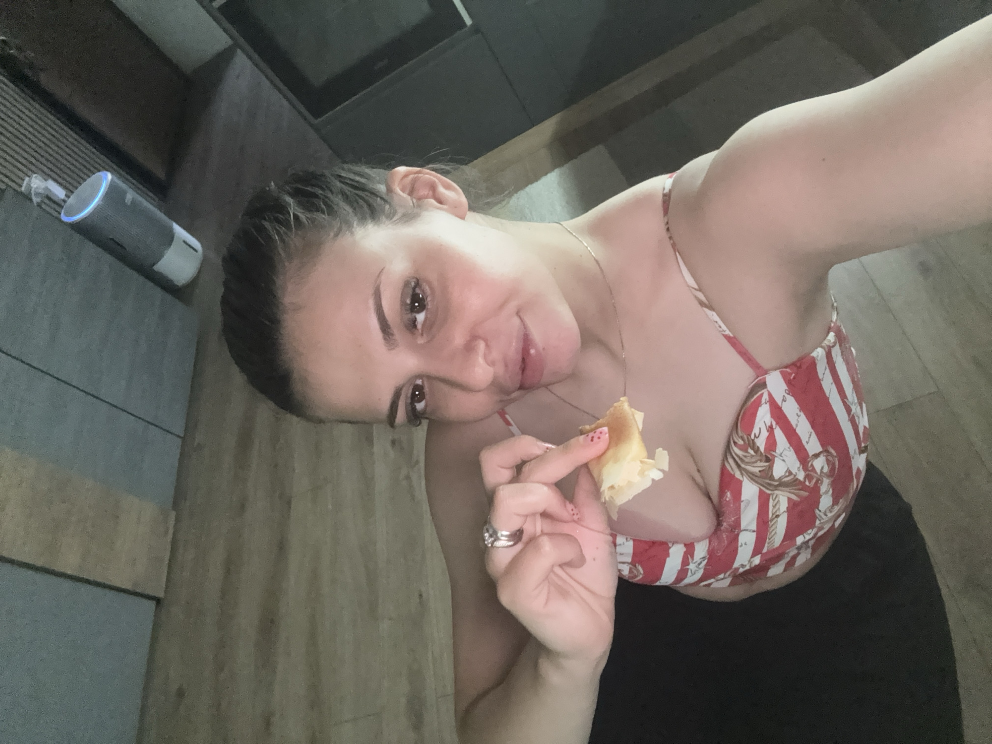 A woman with dark hair pulled back is holding a piece of food up to her face. She is wearing a red and white striped top with a nautical theme. The background appears to be indoors, with wooden flooring and a grey tiled wall. A cylindrical smart speaker is visible on the ceiling.
