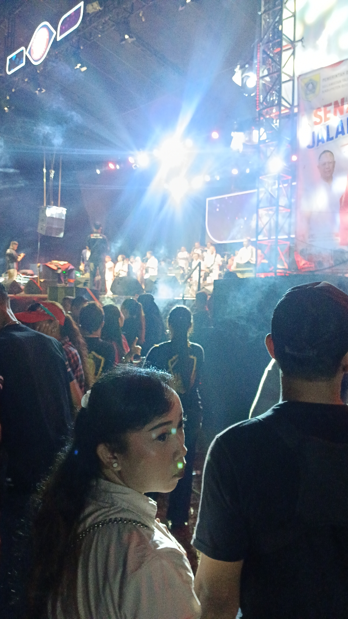 The image shows a crowded outdoor event, likely a concert or festival, with a bright stage illuminated by numerous spotlights. In the foreground, a woman with dark hair tied back is looking to her right, her face partially illuminated by the stage lights. She is wearing a light-colored jacket with a decorative chain detail. To her right, the back of a man wearing a dark t-shirt and a dark cap is visible.
In the mid-ground, a diverse crowd of people is gathered, many looking towards the stage. Some individuals are wearing dark clothing, while others are in lighter attire. Behind the crowd, on the stage, performers can be seen, some in white outfits. There are also speakers and lighting equipment.
A large banner is visible to the right of the stage, featuring text that appears to be in Indonesian. The overall environment suggests a lively public gathering in an urban or semi-urban setting, possibly at an event venue or a public square. Based on the provided location information, this scene is situated in Tajurhalang, Indonesia. The atmosphere is energetic and illuminated by the spectacle of the performance.