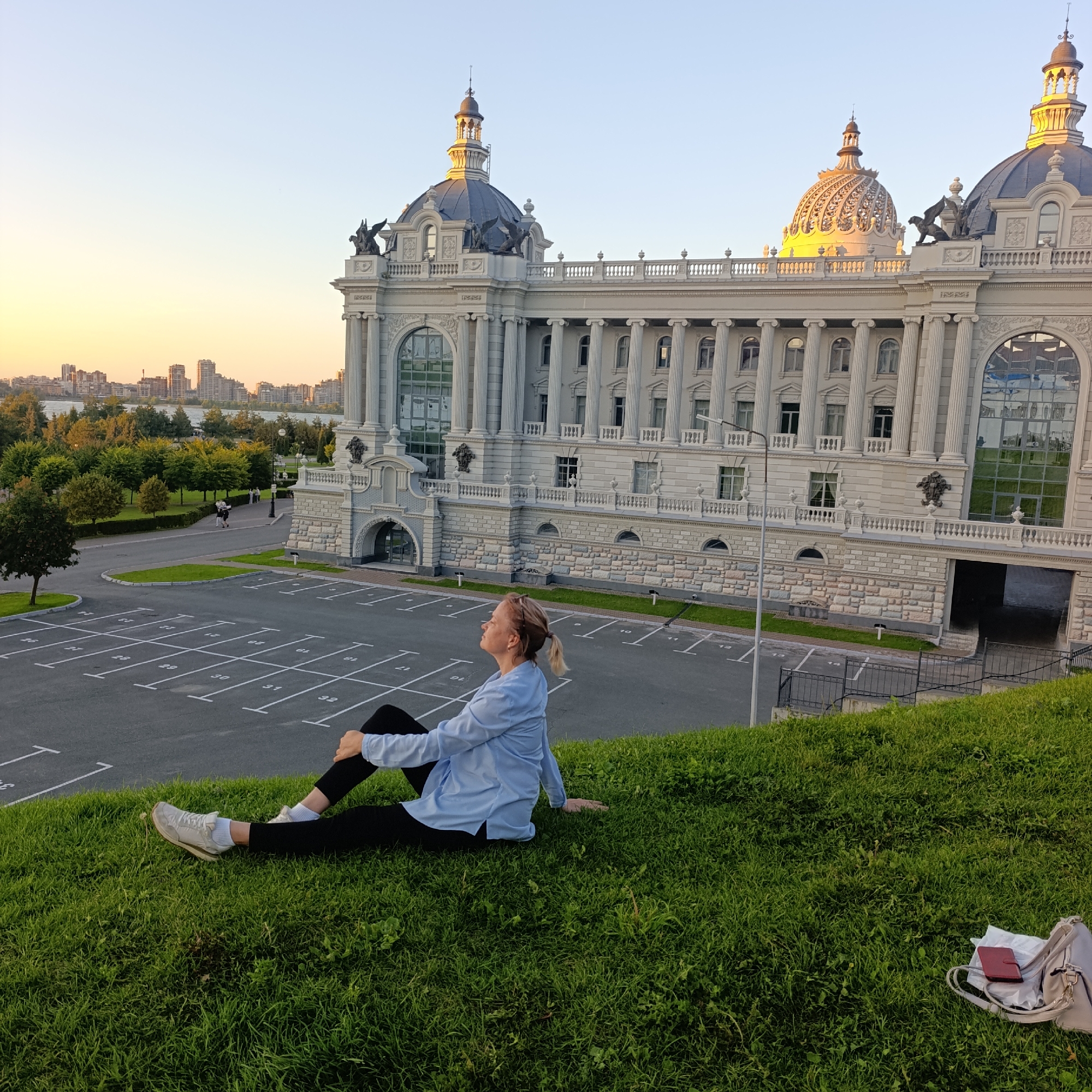 The image depicts a woman sitting on a grassy hill with a large, ornate building in the background. The building, identified as the Palace of Farmers in Kazan, Russia, features classical architecture with columns, domes, and intricate sculptures. The woman is dressed in a blue shirt and black pants, with her legs crossed and her head tilted back, gazing upwards. A light-colored bag and a red wallet are placed on the grass near her. The scene is bathed in the warm light of sunset, casting long shadows across the parking lot in front of the building. In the distance, a city skyline with numerous buildings and a body of water can be seen. The overall impression is one of peaceful contemplation amidst an impressive architectural setting.