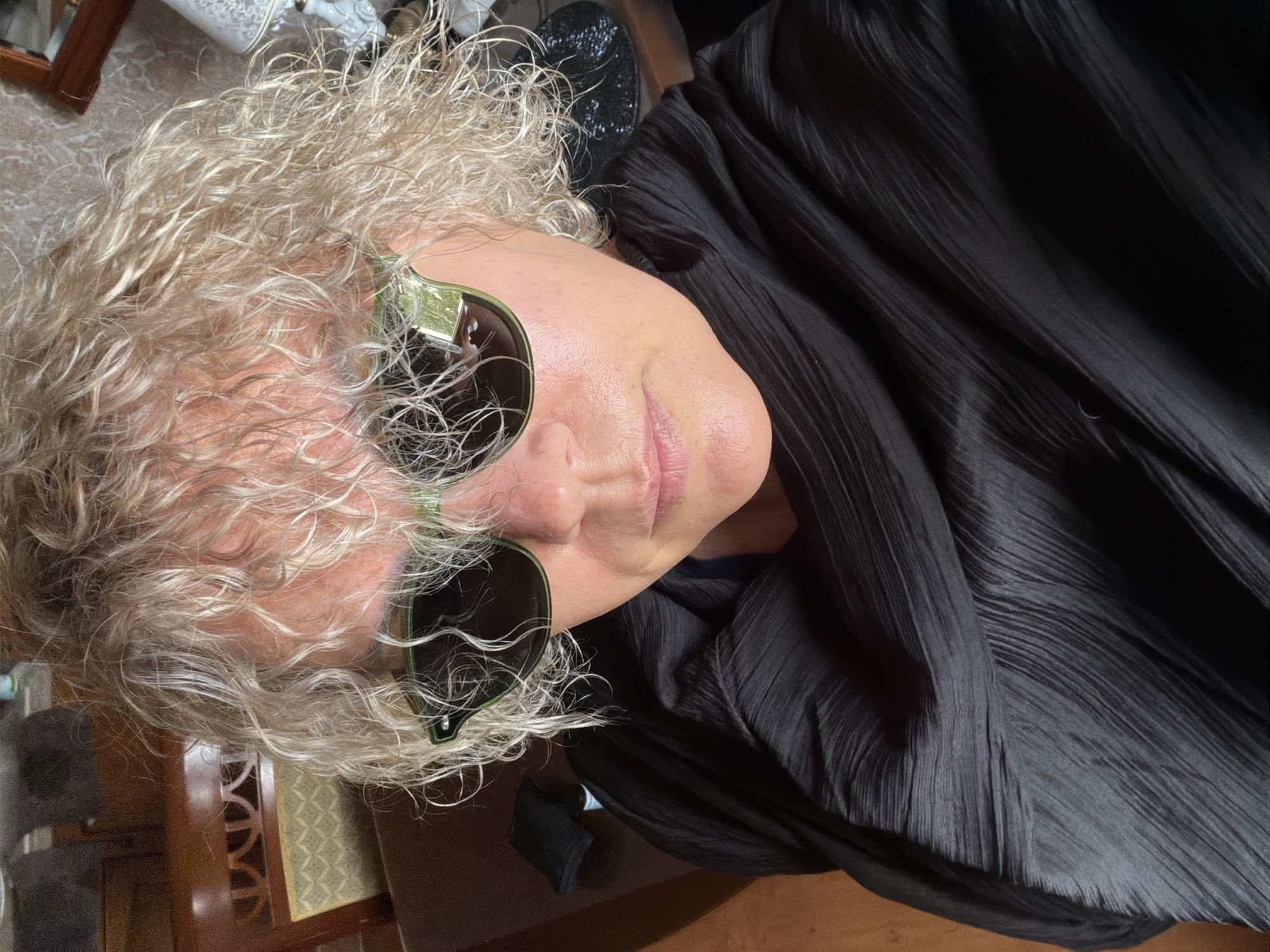 A high-angle, medium close-up shot shows a person wearing sunglasses and a black shirt. The person has curly, blonde hair. The background is out of focus and shows a table and chairs. The lighting is natural and appears to be coming from a window.
