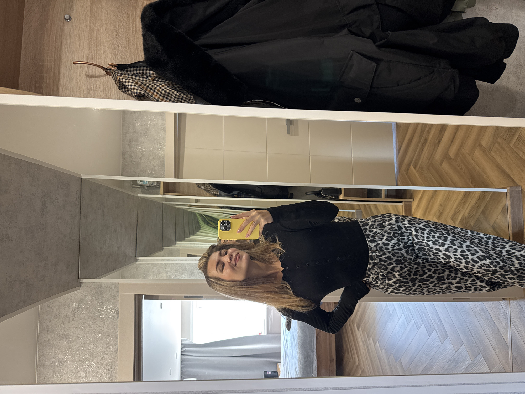 The image is rotated 90 degrees counter-clockwise, depicting a young woman taking a mirror selfie. She has light brown hair and is smiling subtly, holding a bright yellow smartphone with a visible camera in her right hand. She is wearing a long-sleeved black top with buttons down the front and high-waisted leopard-print trousers.
The background reflected in the mirror shows an indoor space, likely a bedroom or dressing area, with a modern aesthetic. Elements visible in the reflection include light-colored, possibly tiled, walls and a light wooden floor with a distinct herringbone pattern. Through an open doorway, a bed with a grey duvet and headboard is partially visible, suggesting natural light is entering from a window. The mirror itself creates a "hall of mirrors" effect, reflecting multiple receding images.
Above the mirror, in the actual foreground of the unreflected room, a dark winter coat with a fur-lined collar or hood is draped, alongside another patterned jacket (possibly plaid) hanging on a wooden hanger. A small, circular, silver-colored metal knob is also visible on a light wood-paneled wall. The scene appears to be taking place during the daytime. No visible text is present in the image.