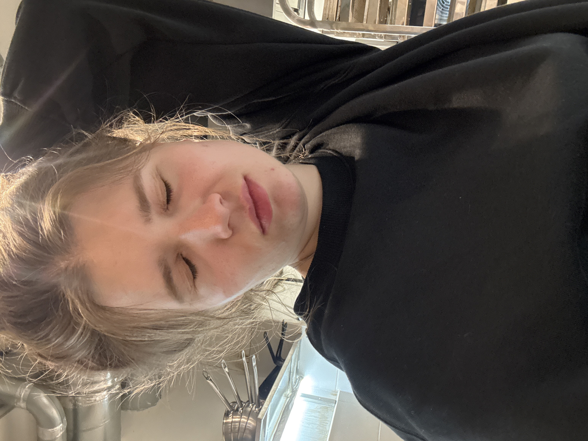 A person with light skin and closed eyes wearing a black hooded sweatshirt is the main subject of the image. The setting appears to be indoors, possibly a kitchen, based on the presence of a utensil holder with various metal utensils and a stainless steel pipe visible in the background. The image seems to be taken from a slightly downward angle. There is no information available in the image to suggest the location is Belgrade, Serbia.