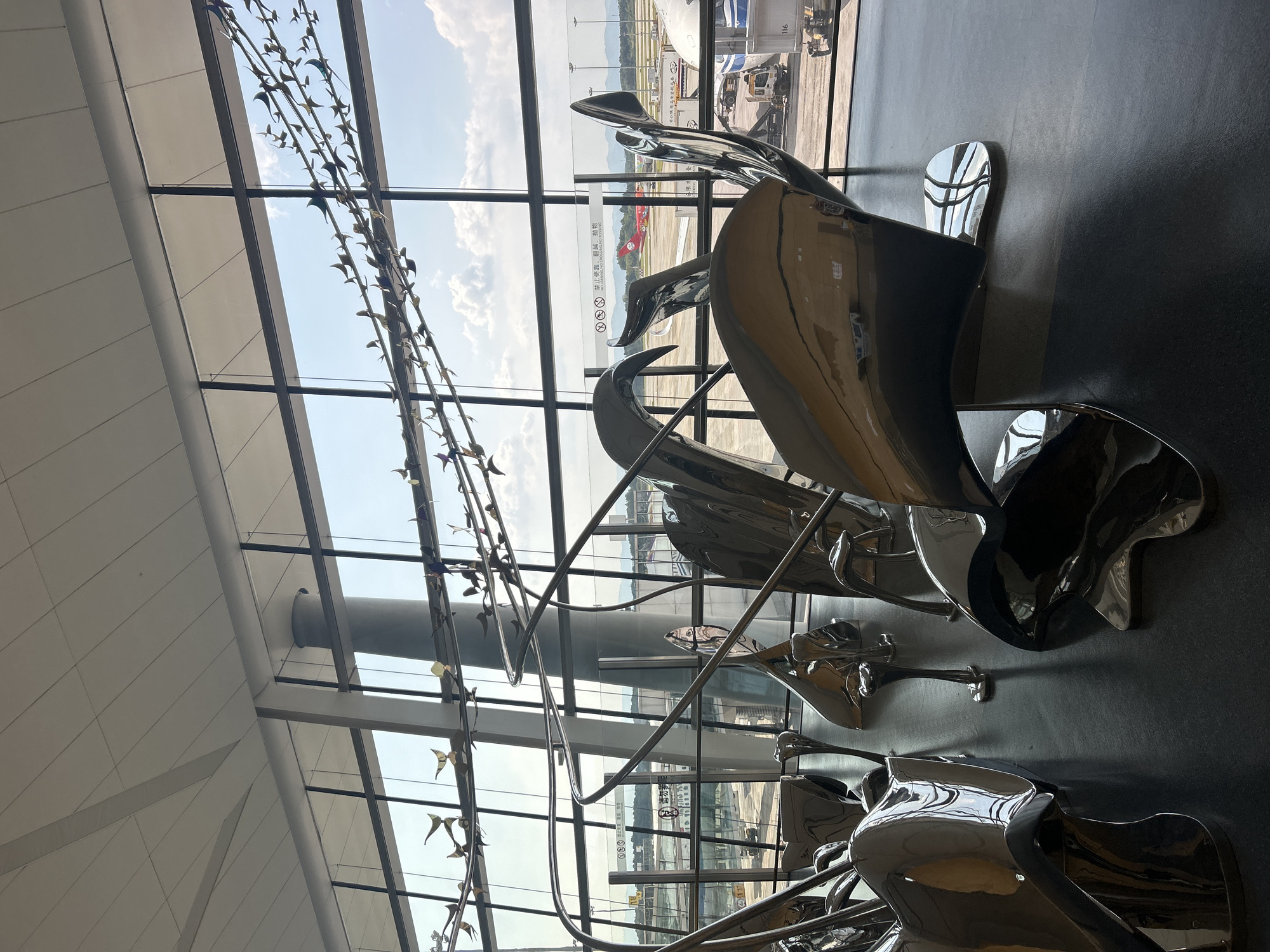 This image depicts the interior of an airport terminal, likely located in Jianyang City, China, given the presence of Chinese signage visible through the large windows. The foreground is dominated by a large, abstract, highly polished metal sculpture. The sculpture has smooth, flowing lines and appears to be composed of multiple interconnected elements, with reflections of the surrounding environment visible on its surface. Behind the sculpture, a expansive window offers a view of an airport tarmac. Several airplanes are visible on the tarmac, along with airport vehicles and structures. In the distance, rolling hills are visible under a bright, cloudy sky. Above the sculpture, a decorative installation of numerous small, metallic bird-like figures is suspended, appearing to be in flight. The floor of the terminal is a dark, reflective material, further contributing to the visual complexity of the scene through its reflections of the sculpture and overhead lighting. The overall impression is one of a modern, spacious, and art-filled public space within an airport.