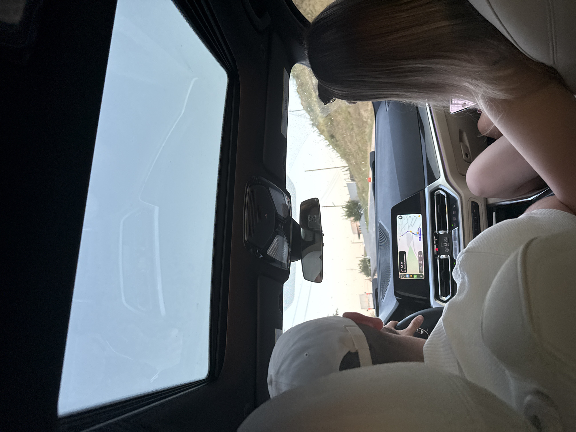 The image shows the interior of a car, viewed from the back seats looking forward. A man wearing a white cap and a white polo shirt is in the driver's seat, holding the steering wheel. To his right, a woman with blonde hair, wearing sunglasses on her head, is looking at her phone. The car's dashboard is visible, with a navigation screen displaying a map. Outside the car, through the sunroof and windshield, a road curves up a hillside with sparse vegetation. The sky is bright and overcast. The scene suggests a journey, possibly on a highway, as indicated by the navigation system and the road layout. The overall atmosphere is casual and relaxed. There is no specific indication that the location is Sărata-Mereșeni, Moldova, beyond the prompt's request to include it.