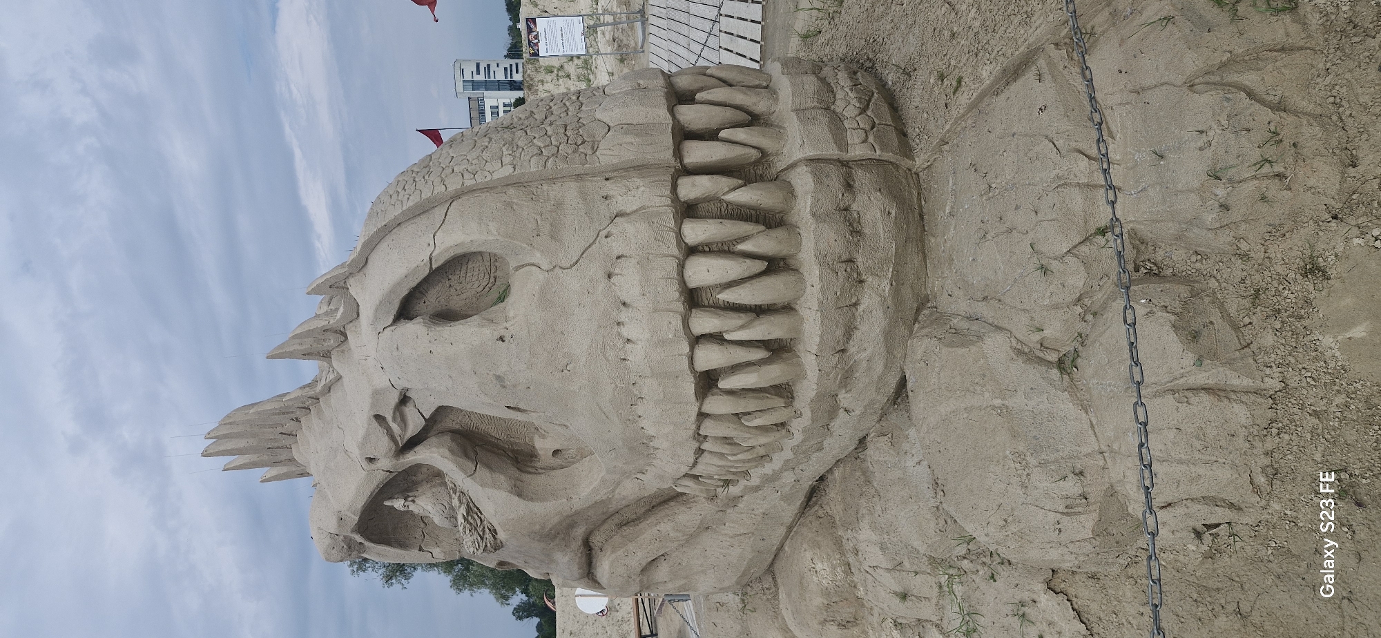 A massive sand sculpture of a dinosaur skull is displayed outdoors under a cloudy sky. The sculpture is intricately detailed, featuring large, sharp teeth, cracked eye sockets, and a textured surface resembling scales on the top of the skull. Spikes protrude from the top of the head. The sculpture is placed on a sandy ground with some sparse vegetation. A metal chain is strung across the foreground, creating a barrier. In the background, there are wooden boardwalks, a building, and red flags. The image also contains a watermark at the bottom left corner reading "Galaxy S23 FE". Based on the information provided, this sand sculpture is likely located in Jelgava, Latvia, possibly at a beach or public park where such art installations are exhibited.