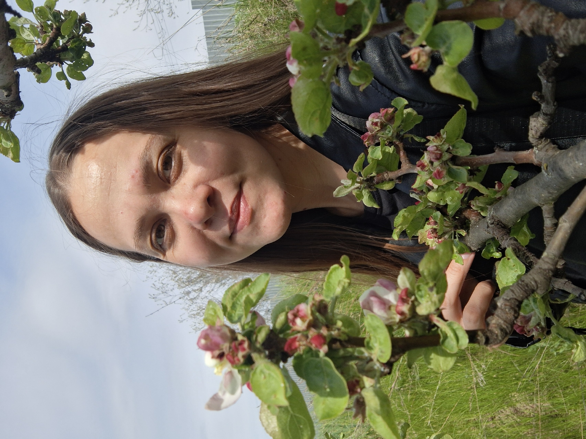 A woman with long brown hair and fair skin, wearing a black leather jacket, stands among blossoming tree branches. The tree branches feature green leaves and reddish-pink buds and blossoms. In the background, there is grass, possibly a field, and a partial view of a light-colored building or structure. The location is presumed to be in Chaikovsky, Russia, given the prompt. The woman appears to be holding one of the branches, possibly interacting with the blossoms. The sky is visible as light blue in the background.