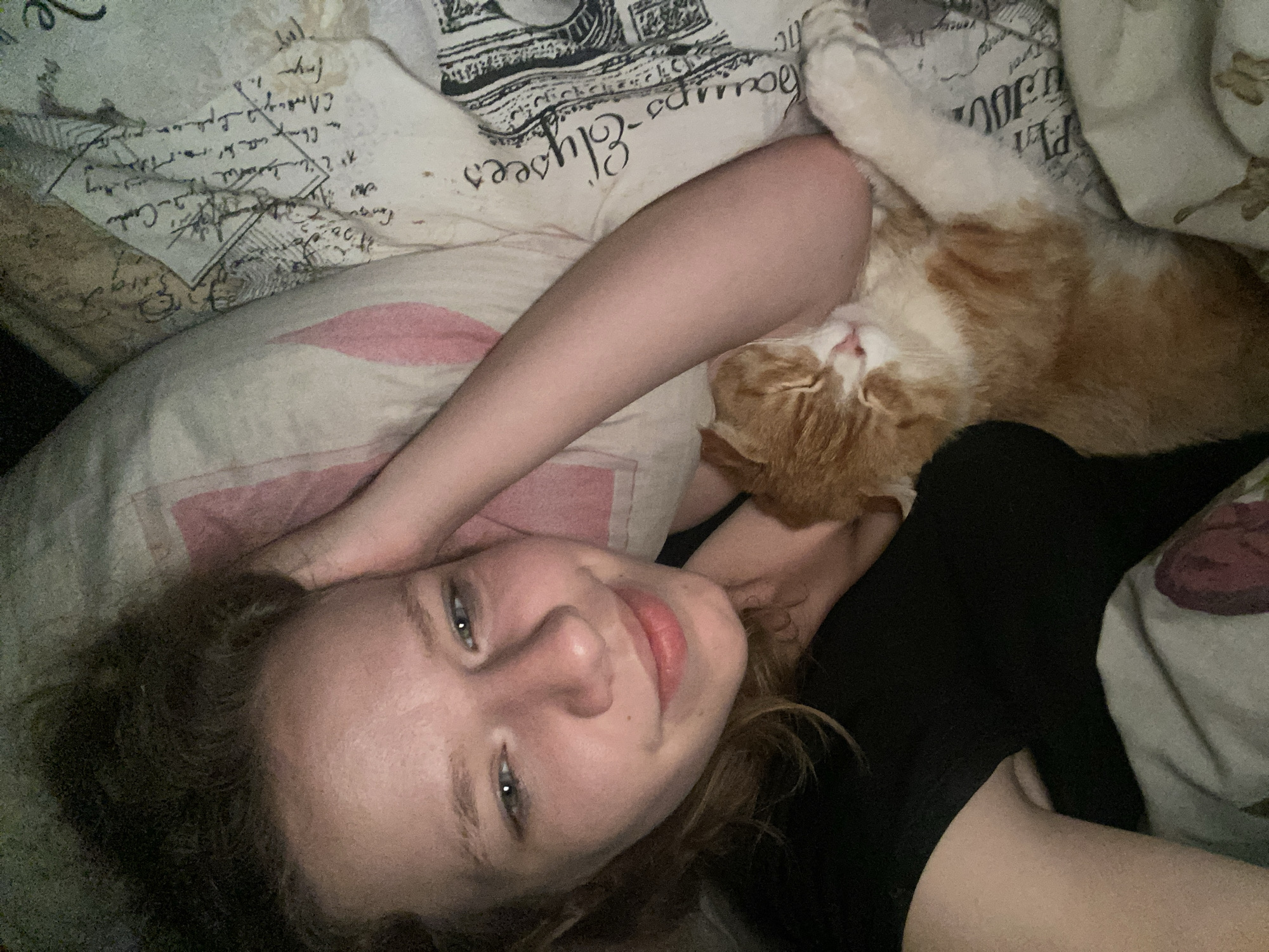 A fair-skinned person with light hair lies in bed, their head resting on a pillow with a faded script pattern, and a pink stripe. They are positioned next to an orange and white cat. The cat is lying on its side, its paws near the person's face. The person's arm is raised and slightly bent, with their hand near their head. The background consists of a bedspread with more script and possible floral pattern. No information or visual cues suggest the location is Pakhynia, Ukraine or any other city.