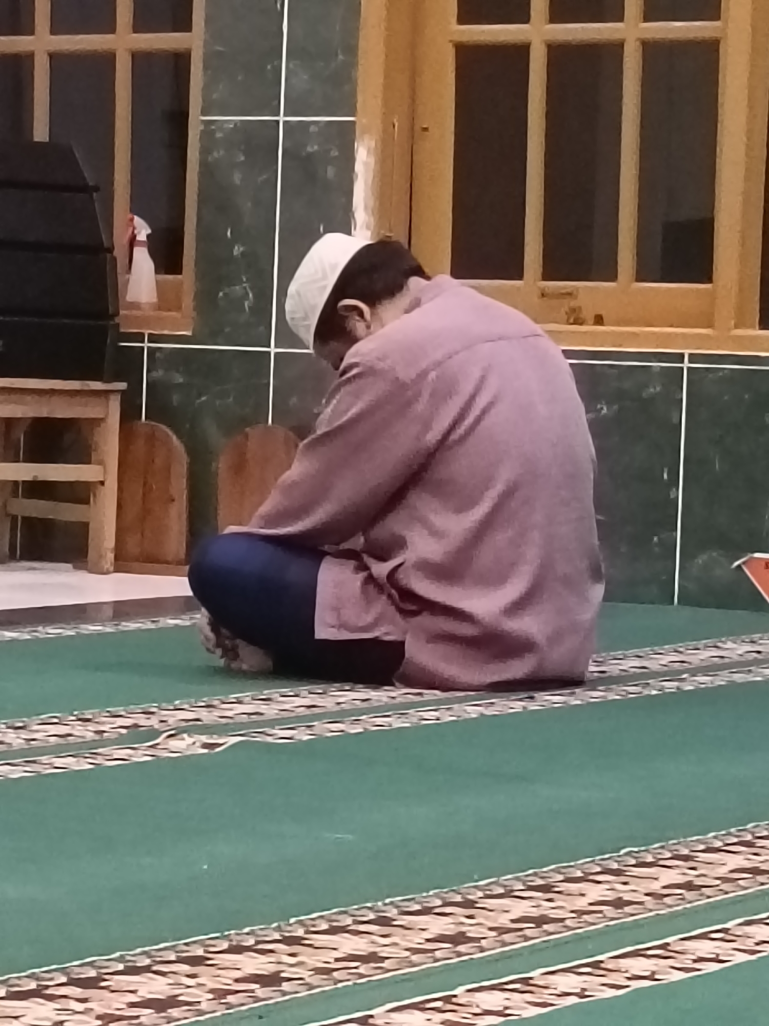A man wearing a white skullcap and a purplish-brown shirt is sitting on a green carpet with a patterned border. He is facing away from the camera, with his head bowed. The man appears to be in a room with dark green tiled walls and a wooden window frame with multiple panes. To the left, there is a wooden table and a dark, rectangular object. The scene suggests an indoor setting, possibly a prayer room or mosque. The location is specified as Kediri City, Indonesia.