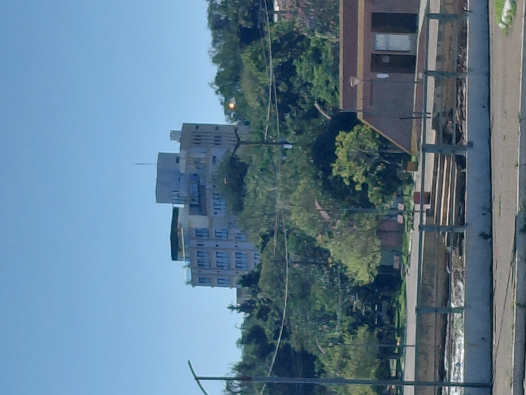 The image shows a distant view of a multi-story building with a flat roof, possibly an apartment complex or hotel, situated on a hill amidst dense green trees. A clear blue sky is visible above. In the foreground, there is a body of water with a concrete embankment and a railing. Further in the foreground, a street or paved area is visible, with some grass on the right side. The building in the background appears to be light-colored with many windows. A streetlamp is visible on a pole to the left of the building. To the right of the building, there is a smaller, dark-colored structure with a pitched roof and windows. The scene suggests an urban or semi-urban environment with a waterfront. This location is likely along the Parana River in Encarnación, Paraguay, given the presence of waterfront infrastructure and the architectural style which is common in the region. No people are clearly discernible in the image.