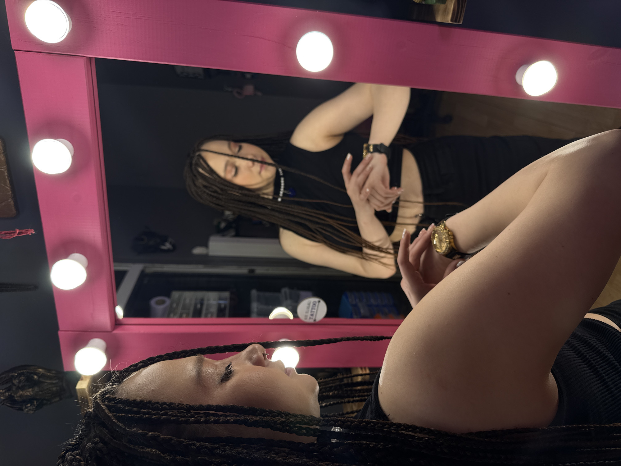 The image depicts an indoor scene within what appears to be a tattoo or beauty studio, located in Saratov, Russia. The central subject is a young female with light skin tone and long, dark micro-braids or dreadlocks. She wears a black sleeveless crop top exposing her midriff, dark pants, a small septum piercing, and a choker-style necklace with white beads and a central dark pendant. Her fingernails are manicured with a light polish.
She is positioned in front of a large, rectangular mirror framed in a vibrant magenta-pink color. Seven round, bright white LED bulbs are visibly mounted along the mirror's frame, providing illumination.
In the foreground, the subject's left arm is prominently displayed, bent at the elbow, with a gold-colored watch visible on her wrist. Her right hand is positioned over this watch, suggesting interaction or adjustment. Her eyes are lowered, indicating focused attention on her left wrist.
The mirror's reflection shows the subject from a frontal perspective, also looking downwards. In the reflection, her right arm (appearing on the left side of the reflection) is visible, with a black smartwatch on its wrist. Her left hand (appearing on the right side of the reflection) is positioned over the smartwatch, indicating interaction or adjustment.
The background visible through the mirror's reflection reveals a room with dark gray or black walls. Key objects include a white circular sign with dark text partially reading "TATTOO," positioned on the right side of the reflection. To the left of this sign, a storage unit with multiple clear drawers containing various supplies is visible. Indistinct dark objects are also present on walls or shelves in the background. Above the mirror in the foreground, a dark, intricately sculpted object, resembling a bust or mask, is mounted on the wall.