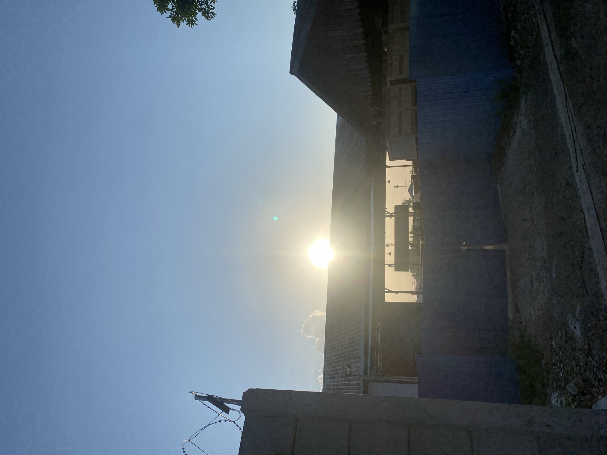 A vertical, eye-level shot shows a bright sun low in the sky, partially obscured by a single, fluffy cloud. The sun is positioned directly above the center of a long, low-lying building with a corrugated metal roof. The building appears to be an open-sided structure, possibly a warehouse or workshop. To the right of this building is a smaller structure with a dark, sloped roof, possibly made of wood. In the foreground, a blue corrugated metal fence stretches across the width of the image, with a dark metal gate in the center. To the left of the fence, a concrete pillar with barbed wire attached to its top juts into the frame. The ground in front of the fence is rough and appears to be dirt and debris. The sky is a clear, pale blue. The scene is likely located in Banjarbaru, Indonesia, given the style of the buildings and the tropical-looking vegetation visible in the distance.