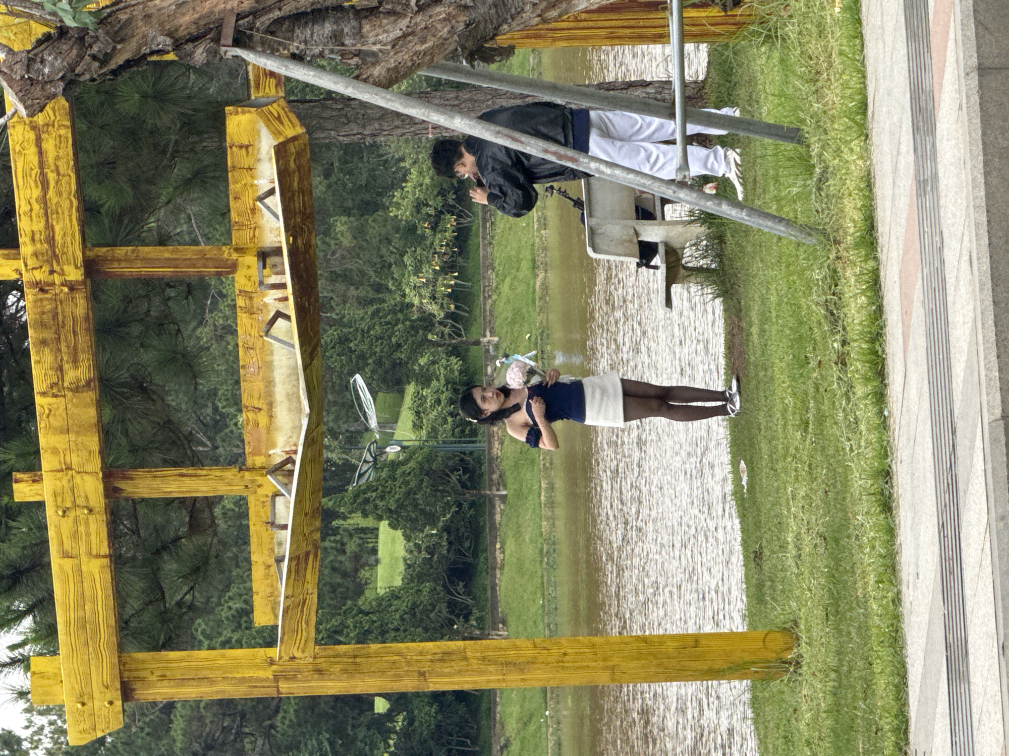 The image depicts a scene in Da Lat, Vietnam, featuring a lake with two people and a yellow, painted wooden structure reminiscent of a torii gate. One individual, dressed in white trousers and a dark jacket, stands on a platform near the lake's edge, appearing to be engaged with a mobile phone, possibly taking a photograph. The second individual, a woman, stands near the water holding a bouquet of flowers, and is wearing a dark top, a white skirt, and dark tights. The lake is surrounded by greenery, including grass and trees. A paved walkway with textured concrete is visible along the right side of the frame. The scene is partially framed by construction scaffolding.