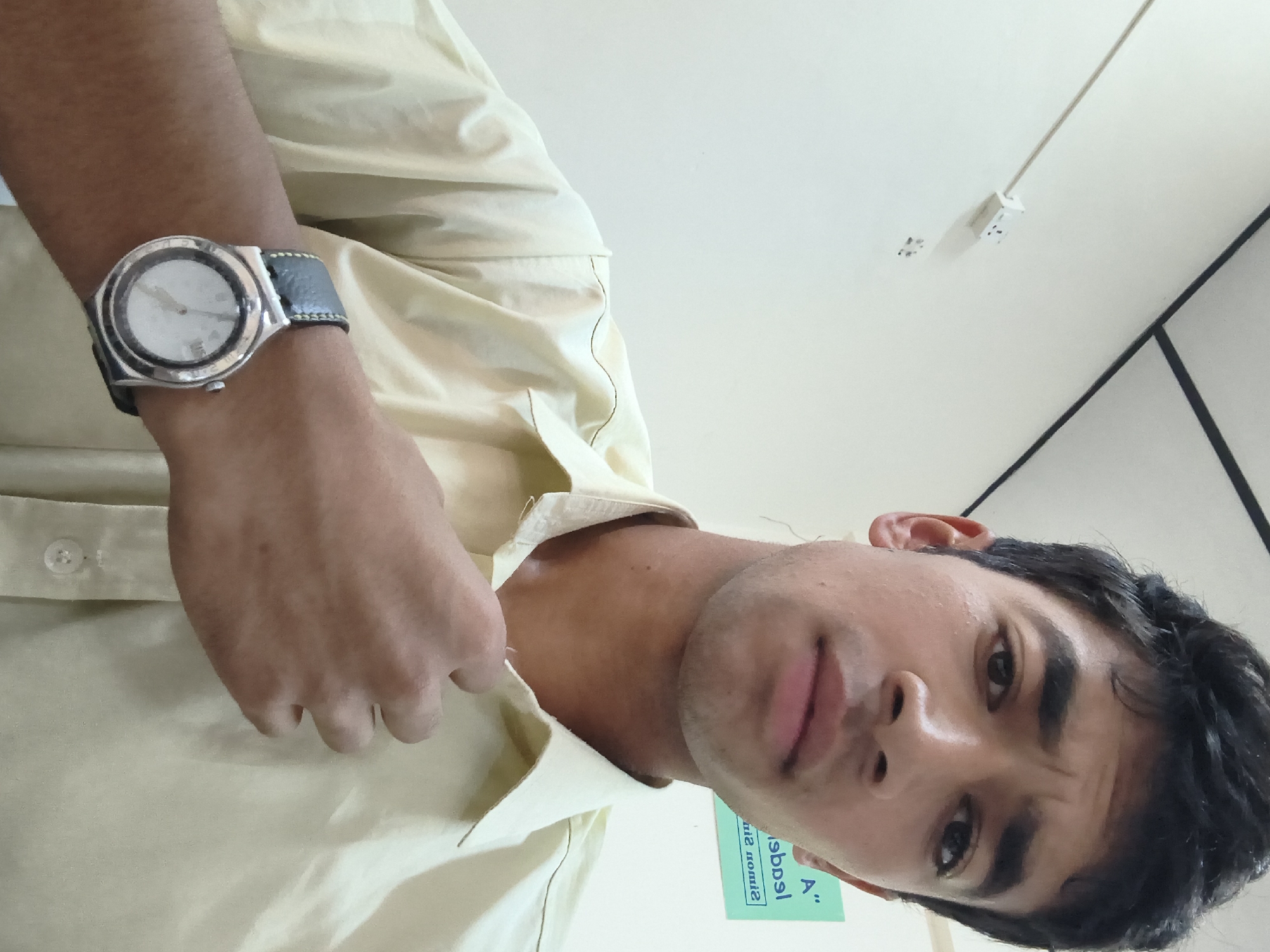 A young South Asian male with dark hair and a light complexion is shown in a close-up, selfie-style portrait. He is wearing a light yellow collared shirt, unbuttoned at the top. His left hand is raised, touching the fabric of his shirt near the collar. A silver wristwatch with a black strap is visible on his left wrist. His eyebrows are furrowed, and he is looking directly at the camera with a serious expression. In the background, a white wall is visible with a power outlet and some writing on a green sign. The ceiling has exposed beams and dark trim. The scene appears to be indoors, possibly an office or classroom environment, located in Dharamshala, India.