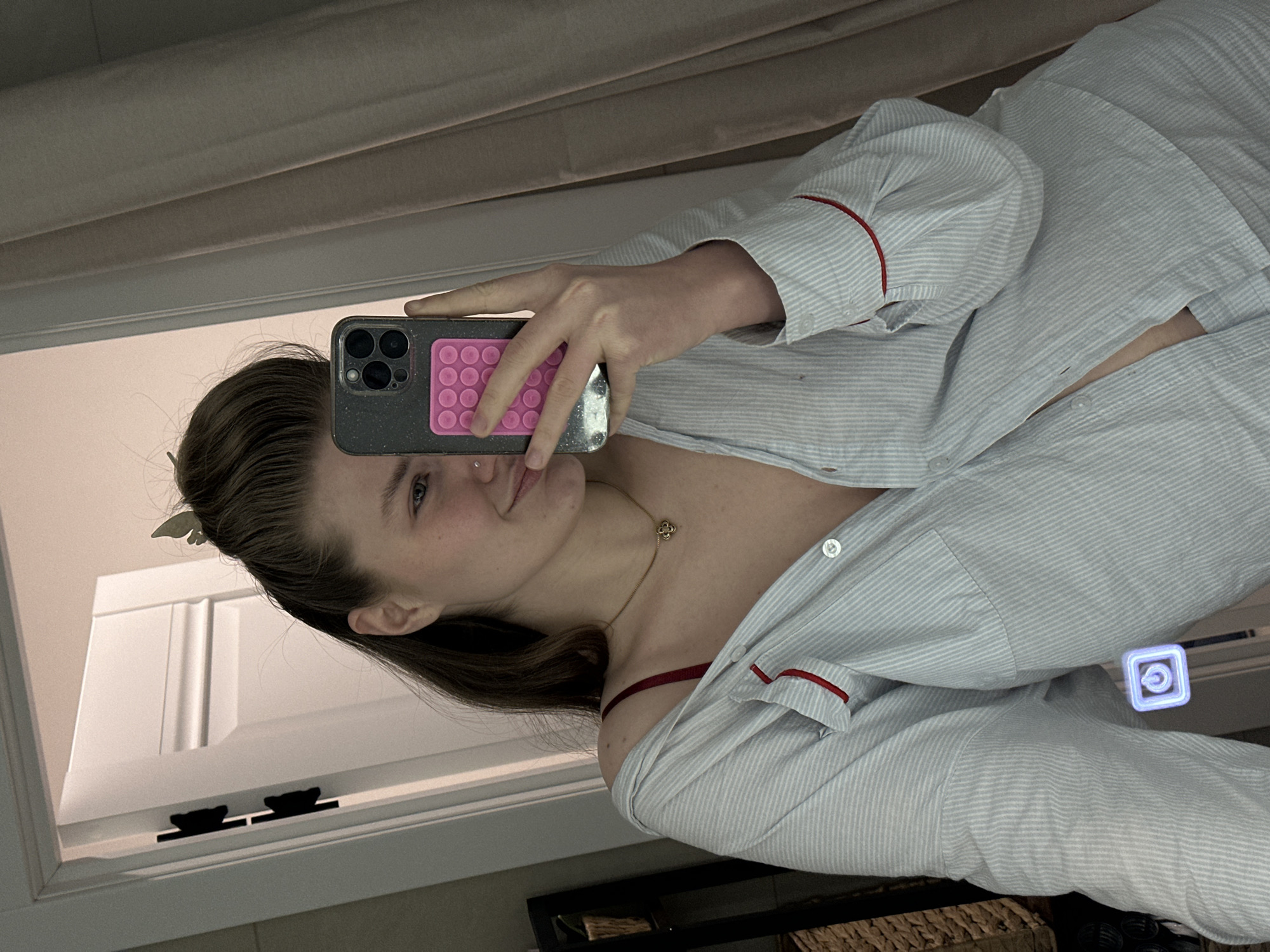 A mirror selfie shows a woman indoors wearing a partially unbuttoned, striped, long-sleeved shirt with red trim. She is holding a phone with a pink accessory on the back. A gold necklace with a clover pendant is visible on her neck. A window with black hardware is behind her on the left. The setting is indeterminate with no indication of the city of Kazan, Russia.
