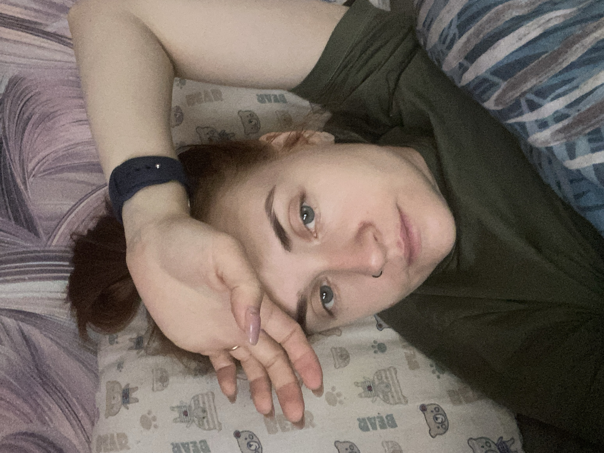A young adult female is depicted lying down indoors, likely in a bedroom setting, with the shot taken from a slightly elevated, angled perspective. She has light skin, reddish-brown hair, and blue-grey eyes that meet the camera's gaze. Her eyebrows are dark and well-defined, and she wears a small ring-style nose piercing on her left nostril. She is dressed in a dark green or olive-colored top.
Her left arm is bent, and her hand is raised, casually resting near her forehead or face. On her left wrist, she wears a dark blue or black smartwatch or fitness tracker, and a small, gold-colored ring is visible on one of her left-hand fingers. Her fingernails show a light, possibly light purple or pinkish, polish.
She is lying on a pillow adorned with a pattern of cartoon bears and paw prints, where the word "BEAR" is clearly visible, appearing both upright and reversed due to the pillow's orientation. The background comprises bed coverings: a solid dark fabric to her right, and a striped bedspread or blanket featuring shades of blue, purple, and white/grey on the left side of the frame. Her expression is neutral, conveying a sense of relaxation or mild contemplation. The scene captures a quiet moment of rest. The location context for this image is Chaikovsky, Russia.