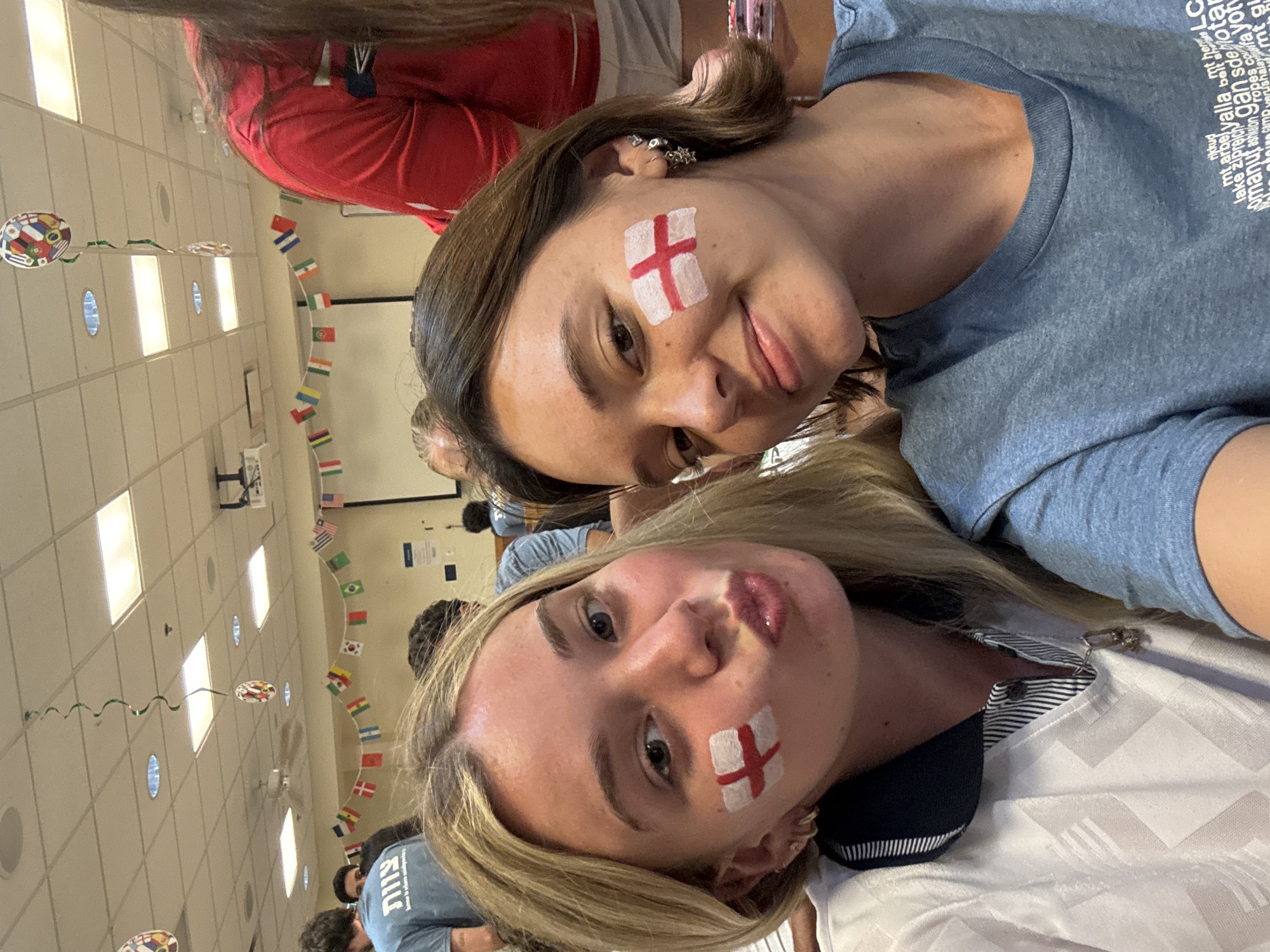 Two young women, with England flags painted on their cheeks, pose for a selfie in an indoor room decorated with flags of various countries. The woman on the left has blonde hair and is wearing a white and navy collared shirt. She is puckering her lips. The woman on the right has brown hair and is wearing a blue t-shirt. She is smiling. In the background, other people are visible, as well as a projector screen and ceiling lights. The room appears to be a common area, possibly a school or community center. The image was taken in Washington Township, United States of America.