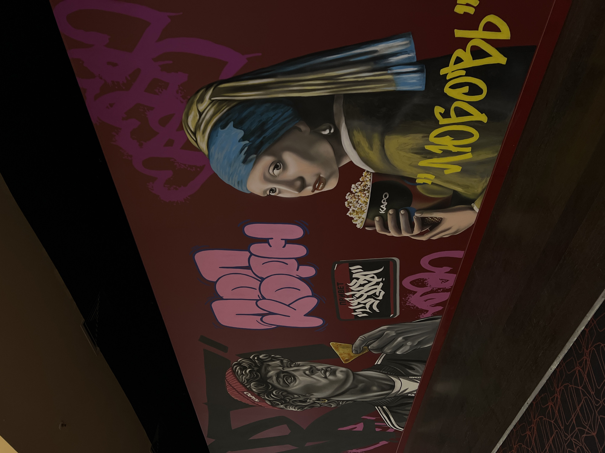 The image shows a wall mural inside what appears to be a commercial space in Khimki, Russia. The mural is a modern artistic fusion, combining classical art imagery with contemporary graffiti elements.
The upper portion of the mural depicts a rendition of Johannes Vermeer's "Girl with a Pearl Earring" holding a bucket of popcorn. In the lower portion, there is a depiction of a bust of Michelangelo's David wearing a beanie and holding a tortilla chip.
Pink and black graffiti-style words are interspersed around the figures. The word "KAPO" is printed on the popcorn bucket. The Russian word "Любовь" (Lyubov, Love) is present near the "Girl with a Pearl Earring" figure. The word "Привет" (Privet, Hello) is near the Michelangelo's David.