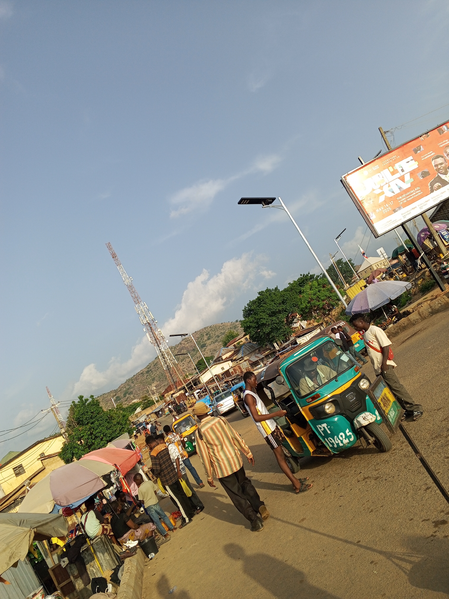 The image shows a bustling street scene in Gwong, Nigeria. In the foreground, a three-wheeled vehicle, a "keke" or auto-rickshaw, is the primary focus. Its license plate reads "PT-19425." Two men stand near it. One, in a white tank top and athletic shorts, appears to be interacting with the vehicle. The other, in a white t-shirt and trousers, leans against it. In the middle ground, there are people walking and gathered near shaded stalls with umbrellas, suggestive of a market area. In the background, there is a tall telecommunications tower. Above and to the right, there's a billboard advertising an event "THE REVAYAH, JOS" scheduled for May 14-17, 2026, at "4 Helen Oomwalk Way, Off Old Airport Junction." The sky is bright blue with scattered clouds.