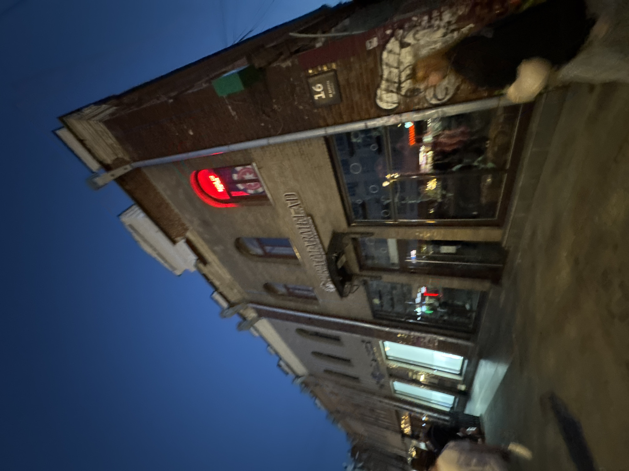 The image depicts a street scene at dusk in Yekaterinburg, Russia. The perspective is angled upwards, focusing on the facade of a brick building. A prominent red neon sign with Cyrillic lettering, appearing to say "ШАР" (SHAR), is visible in an arched window on the upper floor. Below, the building features large glass storefronts with reflections of interior lights and possibly merchandise. A street number sign, "16," is affixed to the brick wall. The sidewalk is paved and reflects the ambient light. Several people are present on the sidewalk, some walking past the storefronts, and one individual with reddish hair is partially visible in the foreground on the right, facing away from the camera. The sky is a deep blue, indicating twilight. The building's architecture suggests an older urban setting. Graffiti is visible on the right side of the building's exterior.
