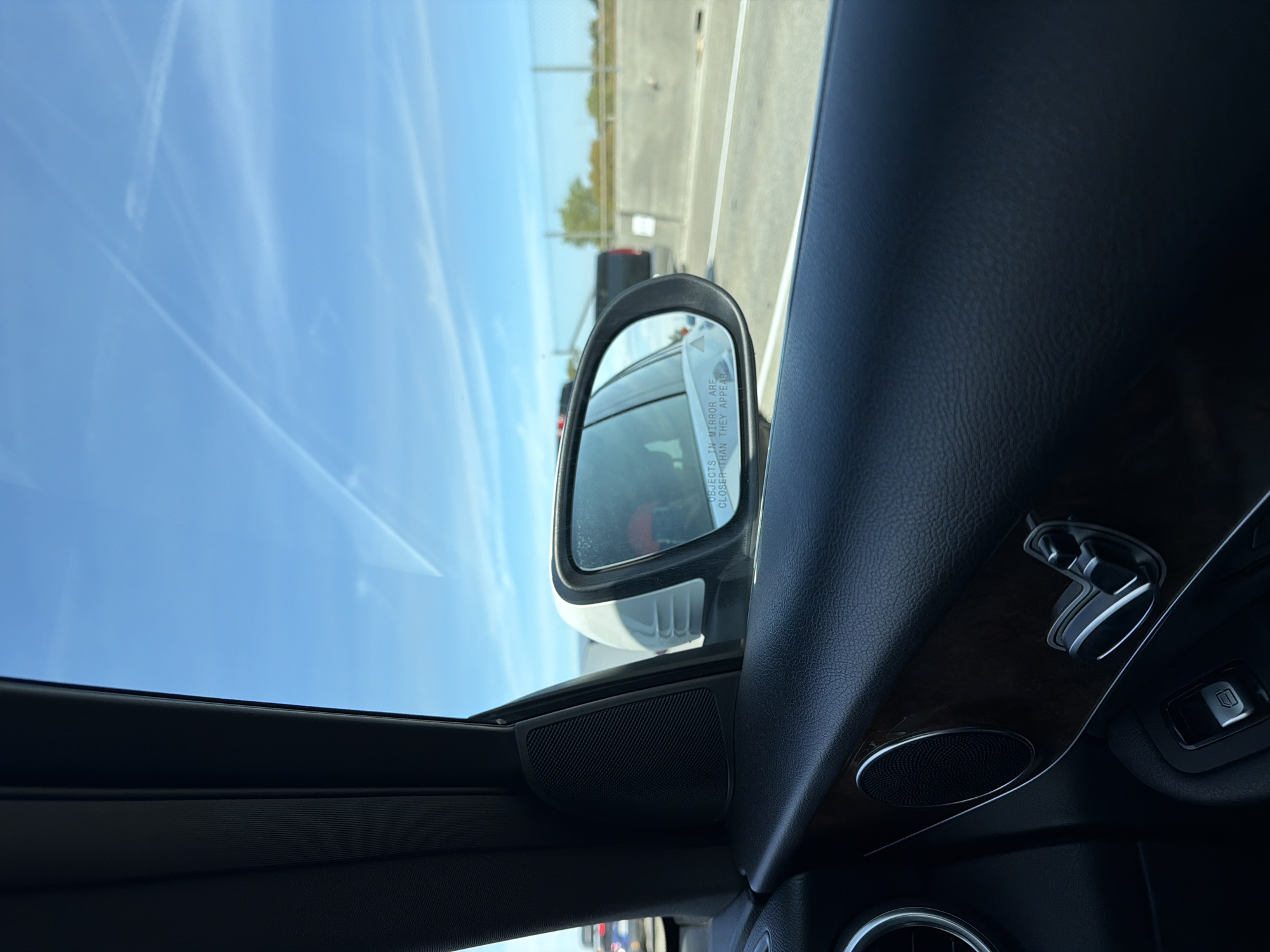 The image, taken from inside a vehicle, captures a clear blue sky with faint contrails, suggesting it's daytime. The foreground features the interior of a car, including a dark leather door panel, a speaker grille, and seat adjustment controls. The side-view mirror is visible, reflecting a white vehicle behind and displaying the warning "OBJECTS IN MIRROR ARE CLOSER THAN THEY APPEAR." The exterior view shows a highway with other vehicles, a concrete barrier, and some trees in the distance, indicating the car is in transit. The setting appears to be a highway in or near West Sacramento, California, United States, given the common highway infrastructure and the presence of typical vegetation. There are no people visible in the frame.