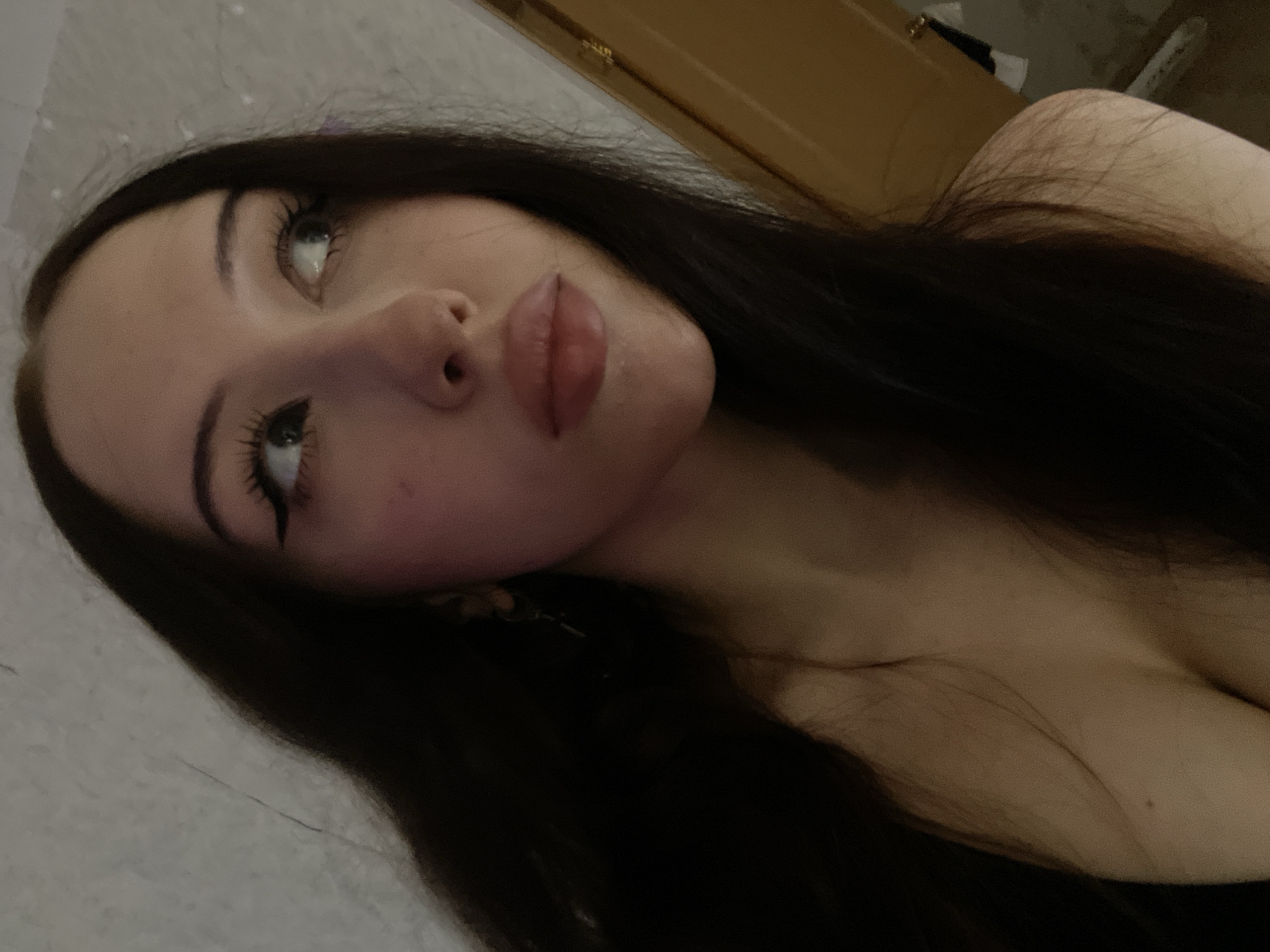 The image is a close-up, indoor, portrait-style photograph of a young woman with long, dark hair. Her head is turned to the right, and she is looking upwards and to the right. She is wearing dark eyeliner and mascara, and her lips are slightly parted and appear to be glossed. Her skin is fair, with some blush on her cheeks. Her cleavage is visible, and she is wearing a dark top or dress. The background appears to be a lightly textured wall and a wooden door frame on the right side of the image. There is no indication of the specific location within Votkinsk, Russia.