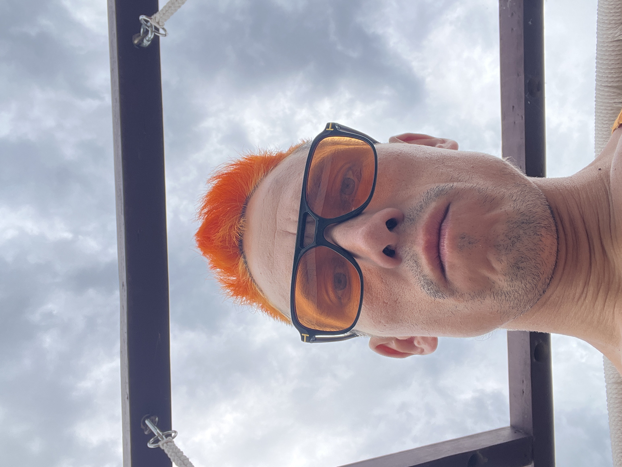 A low-angle, outdoor portrait shows a person with orange hair and sunglasses standing beneath a structure comprised of dark metal poles and white rope. The person's face is centered, with the sunglasses reflecting the sky and obscuring the eyes. The cloudy sky fills the background. No details indicate the location is Chaikovsky, Russia.