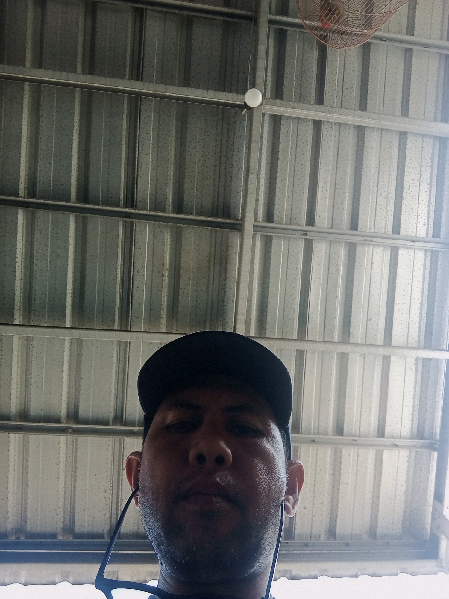 The image shows a man wearing a black baseball cap and sunglasses, looking up at the camera. He is standing under a corrugated metal roof. There is a ceiling fan visible in the background. The scene appears to be outdoors, possibly in a public space. The lighting suggests it is daytime. The image does not contain any information that can determine the specific location within Ketapang Lor, Indonesia.