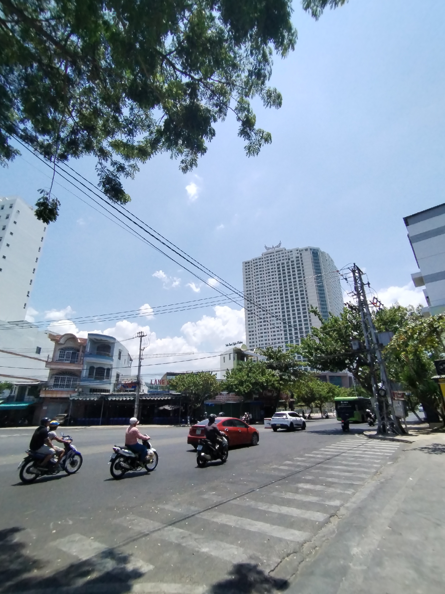 The image depicts a street scene in Nha Trang Ward, Vietnam, under a bright, sunny sky with scattered clouds. A tall, white hotel building with a distinctive emblem on top dominates the background. Three people are riding motorcycles on the left side of the image, crossing a zebra-striped crosswalk. A red sedan and a white car are visible in the background, along with a green bus. Overhead power lines run across the sky. Trees are present both in the foreground and along the street. Other buildings are also visible along the street.