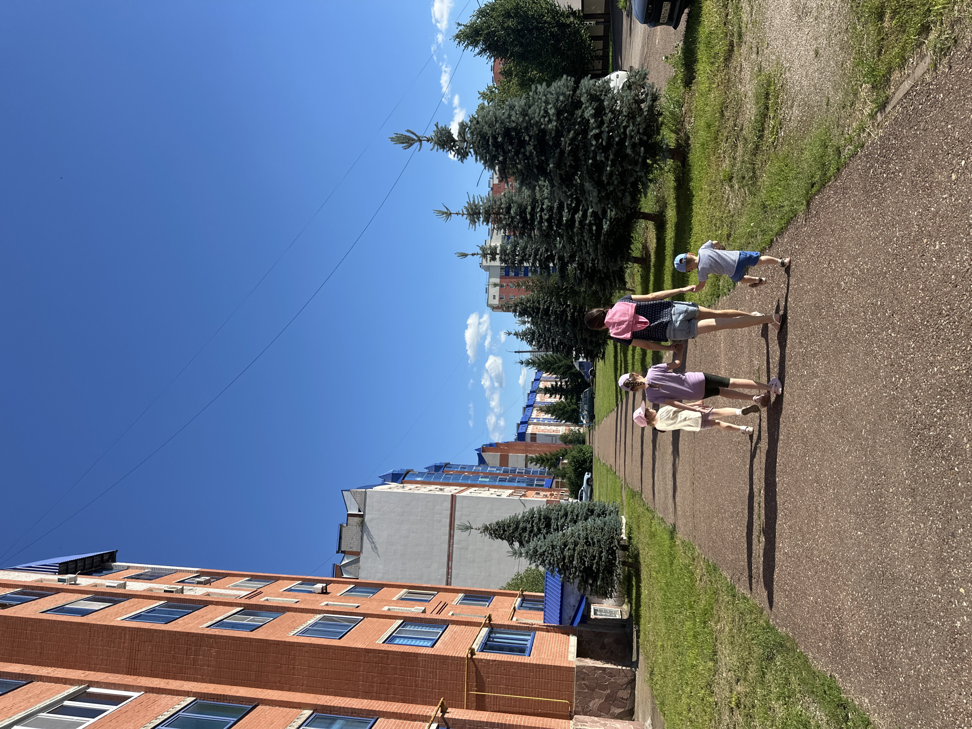 The image shows three young people walking on a paved path next to a grassy area. The person in front is wearing a pink shirt and dark shorts, with a pink backpack. To their right, another person in a purple shirt and dark shorts walks alongside. A third person, wearing a striped shirt, blue shorts, and a blue cap, walks slightly ahead of them. All three appear to be children or young teenagers.
Behind the grassy area, several tall evergreen trees are visible, casting long shadows on the path. Beyond the trees are residential buildings, some made of red brick with many windows, and others with lighter facades and blue-tinted windows. The sky is a clear, bright blue with a few white clouds. The sun is high in the sky, indicating it is likely midday. The overall scene suggests a warm, sunny day in a residential area, possibly a park or courtyard. There are no visible emotions conveyed, but the activity appears to be casual walking or strolling.
The image is oriented vertically, with the sky occupying the upper portion and the ground and buildings in the lower portion. The paved path curves gently to the right as it recedes into the distance. There is a car parked on the side of a road visible in the background, to the left of the evergreen trees. The location context is identified as Mariinsky, Russia. No visible text is present in the image, except for what might be on the license plate of the car in the background, which is too blurry to read.