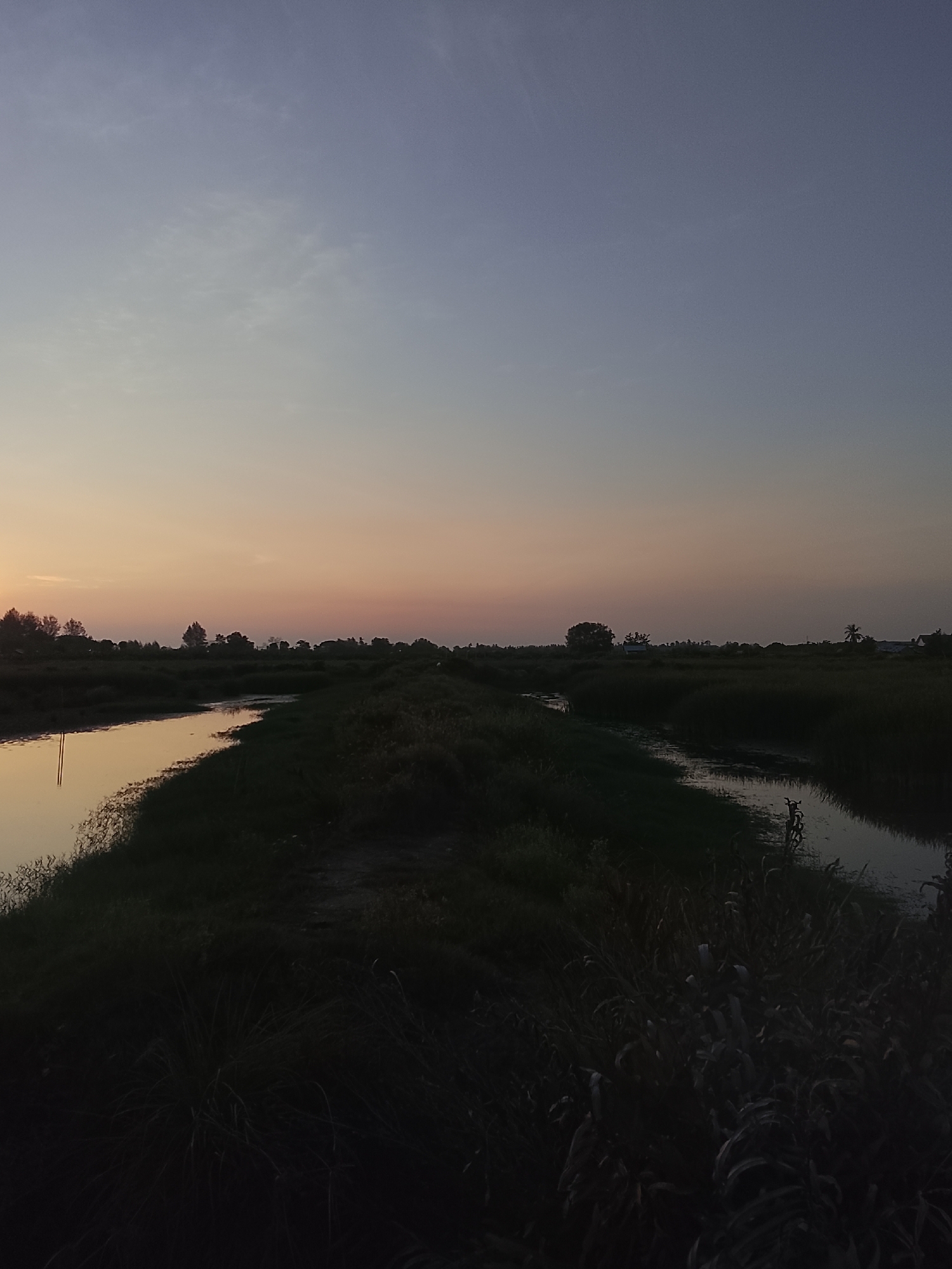 This is a landscape photograph taken at dusk in Mesjid Tuha, Indonesia. The image features a tranquil scene with a body of water reflecting the fading light of the sky. The water appears to be a canal or small river, bordered by dense vegetation on both sides. The sky transitions from a pale orange and pink hue near the horizon to a deep blue at the top of the frame, suggesting the sun has recently set. Silhouetted trees and foliage line the distant horizon, adding depth to the composition. The overall atmosphere is serene and natural.