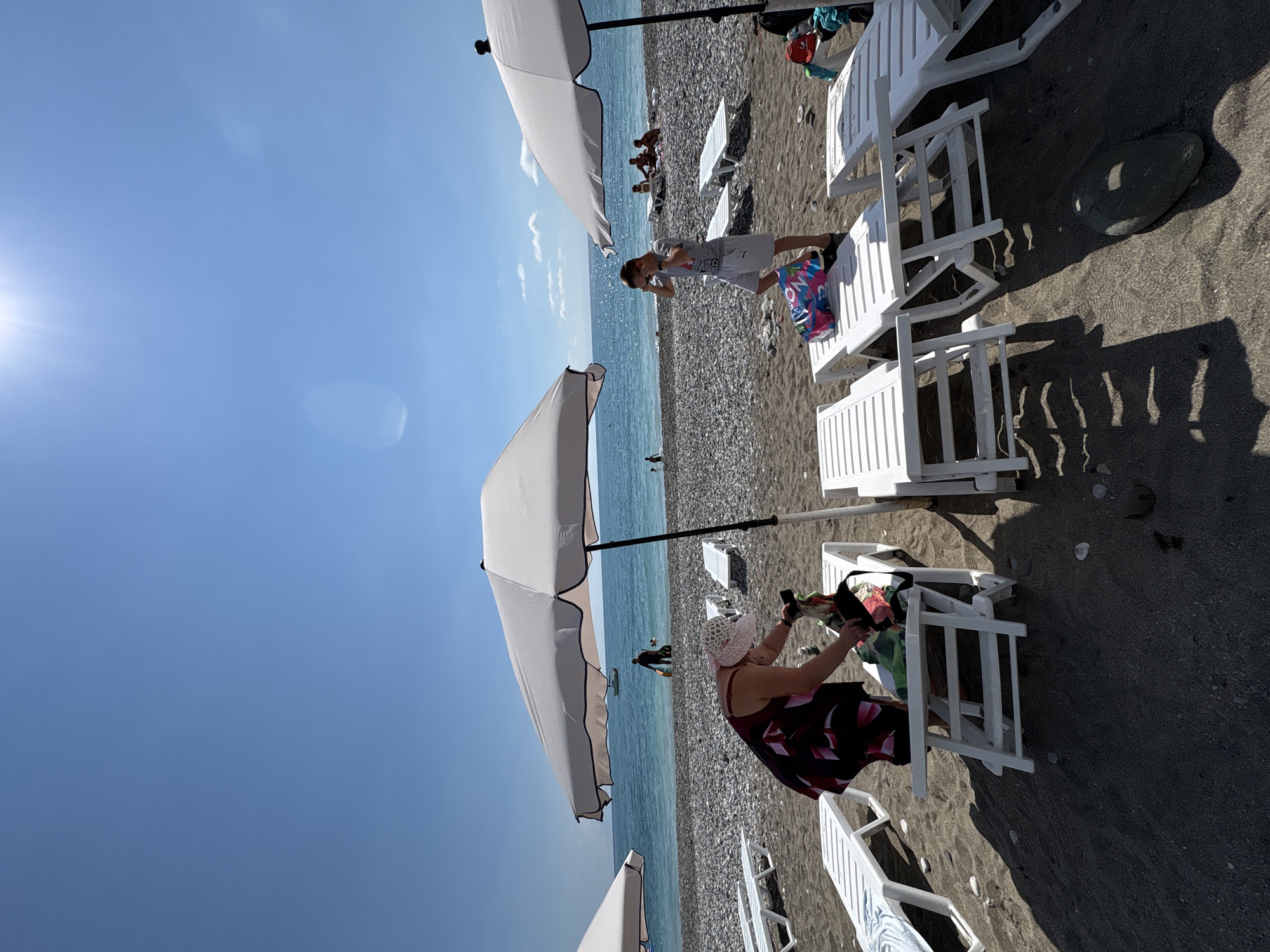 On a sunny day at a beach in Abaata, Abkhazia, several beach chairs and umbrellas are set up on the sand and pebbles. In the foreground, a person in a patterned dress and a wide-brimmed hat is tending to items on a lounge chair. To the right, a young person in a grey t-shirt and shorts stands near a colorful bag on another lounge chair, adjusting their eyewear. In the background, the turquoise ocean stretches out under a clear blue sky, with a few boats and people visible near the water's edge.
