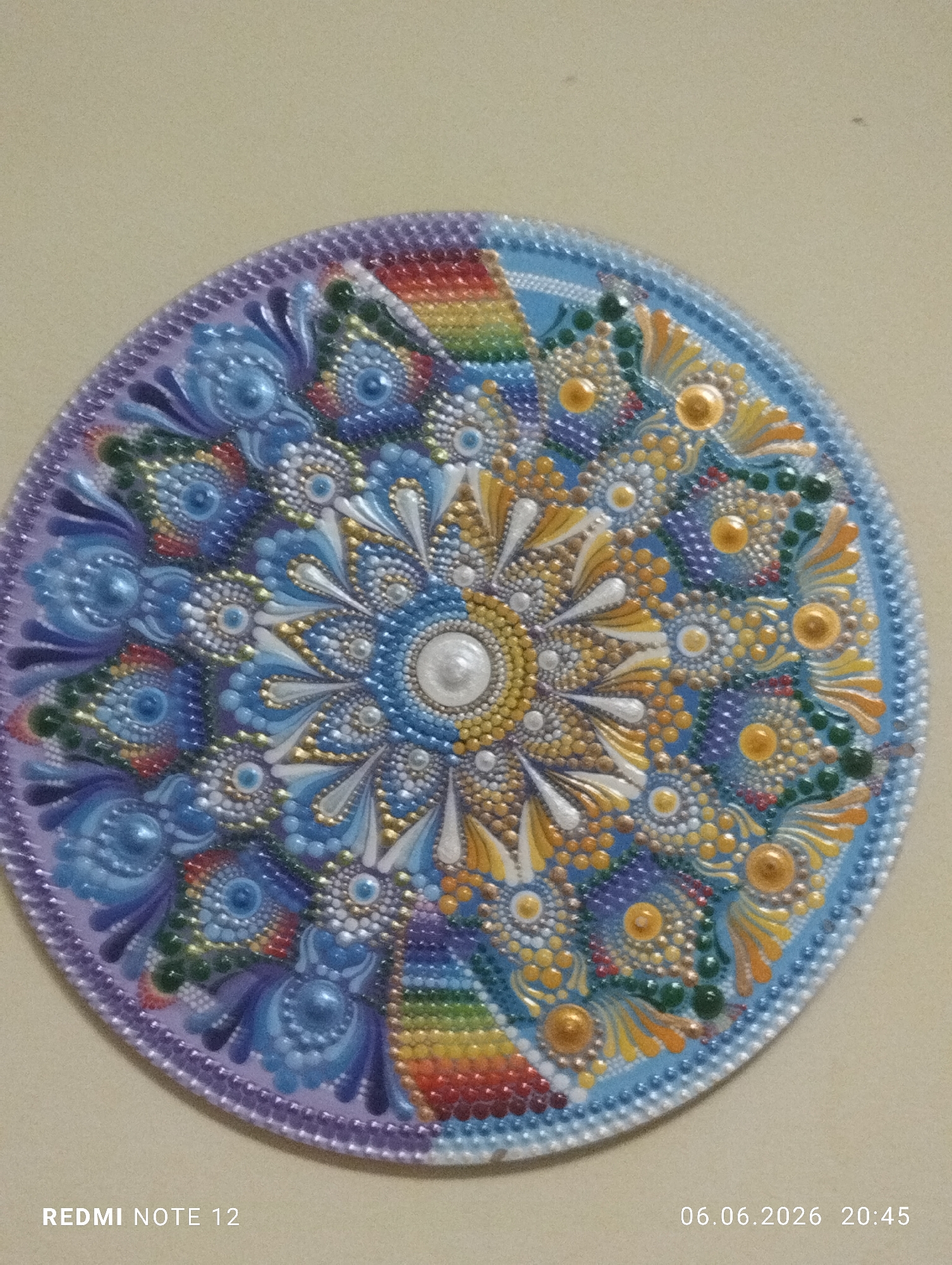 The image displays a circular, three-dimensional artwork, likely a mandala, presented against a plain, light beige wall. The artwork is characterized by an intricate, radially symmetrical pattern composed of numerous raised dots of paint, giving it a textured, tactile appearance.
At the center is a prominent, large, spherical white dot, resembling a pearl. This central element is encircled by concentric rings of smaller dots, primarily in shades of yellow and gold, transitioning to blue and white. Radiating outwards from the center are distinct segments or 'petals' filled with various dot patterns and some smoother painted elements.
The color palette is rich and varied. Blues, ranging from sky blue to deeper indigo, dominate several radial sections and form a significant portion of the outer rings. Yellows and golds are prominent near the center and in alternating radial segments. White and silver dots are interspersed throughout for highlights and specific patterns. Greens appear as dot clusters in some outer sections. The artwork also features segments with a rainbow pattern of dots, showcasing distinct bands of red, orange, yellow, green, and blue. The outermost rim of the circle is bordered by a solid band, purple on the left side and light blue on the right, both adorned with closely spaced, uniform dots along their edges.
The raised dots create a distinct shadow and highlight effect, emphasizing the three-dimensional nature of the painting technique. The background is featureless, providing no environmental context beyond a flat wall.
Overlaid text watermarks are present in the lower corners: "REDMI NOTE 12" in the bottom left, and "06.06.2026 20:45" in the bottom right, indicating the device and timestamp of the photograph. No people, interactions, or discernible environmental features specific to a city or country are present in the scene.