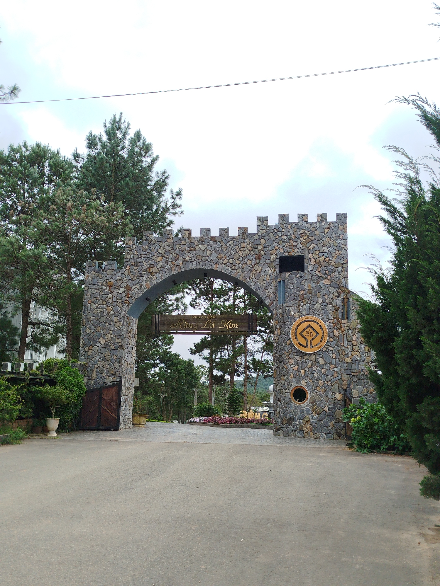 The image shows the arched stone gateway to the tourist area "Khu Du Lịch Rừng Lá Kim" in Lâm Viên - Đà Lạt, Vietnam. The gateway is constructed with rough-hewn stones and features crenellations along its top, resembling a castle. A wooden sign with the text "Khu Du Lịch Rừng Lá Kim" is hung beneath the arch. To the right of the arch, a circular emblem with a mountain and tree motif is embedded in the stone. Wooden double doors are visible to the left of the gateway. The surrounding environment includes pine trees, other lush greenery, and a paved pathway leading through the archway. The sky is overcast.