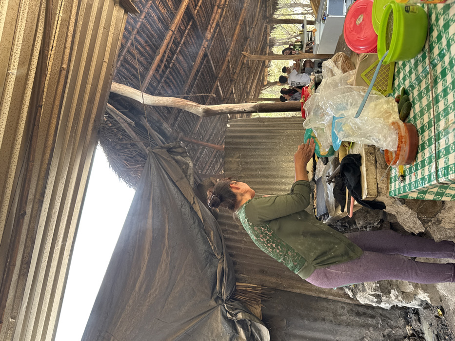 The image depicts an outdoor food preparation and dining area in San Miguel Atlapulco, Mexico. In the foreground, a woman with dark hair tied in a bun, wearing an olive green long-sleeved shirt with an intricate pattern on the back and light purple leggings, stands facing right. Her hands are positioned over a green bowl on a stone countertop, appearing to be shaping dough. A dark griddle and a black cloth are visible on the counter. To her right, a lower table is covered with a green and white checkered tablecloth, holding a terracotta-colored bowl with red salsa, a light blue spoon, a green plastic bowl with green salsa, and several small green vegetables. Multiple clear plastic bags filled with various food items are piled on the counter. The background features a rustic structure with a multi-layered roof system composed of horizontal wooden slats, a dark grey tarp, and a thatched section supported by rough wooden poles. Corrugated metal sheeting forms part of the wall behind the woman. To the far right, several individuals are seated at a white rectangular table, engaged in dining and socializing, with beverage bottles visible on their table. A white rectangular cooler or storage unit is present on the far right.