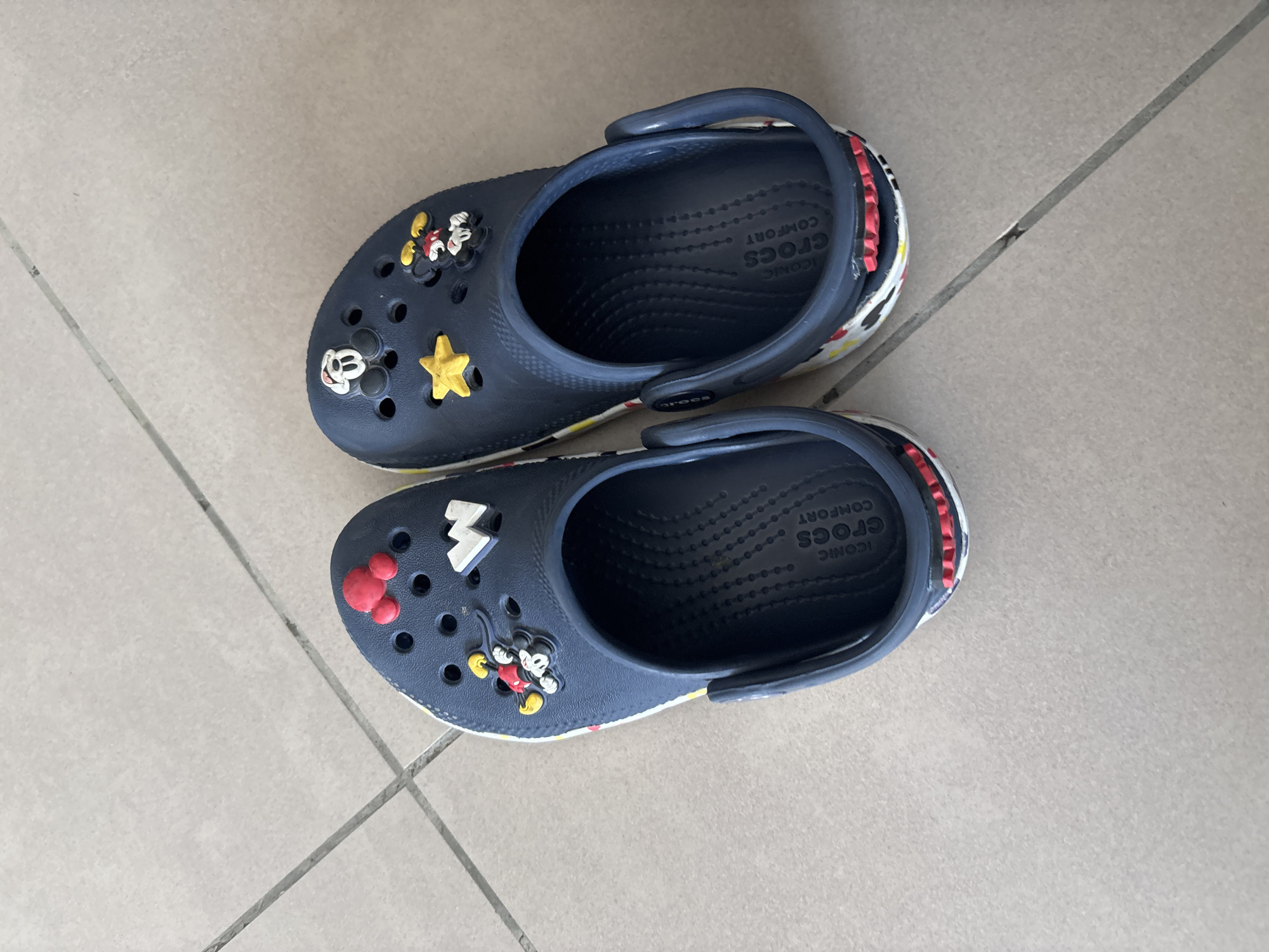 Two dark blue Crocs are displayed on a light-colored tiled floor. The Crocs are decorated with various charms. The left Croc has red Mickey Mouse ears, a yellow star, and a Mickey Mouse charm. The right Croc features a white "W" charm, a yellow star, and a Mickey Mouse charm. Both Crocs have the "ICONIC CROCS COMFORT" logo printed on the inside heel. The tiled floor has visible grout lines. This image was captured in Dubai, United Arab Emirates.